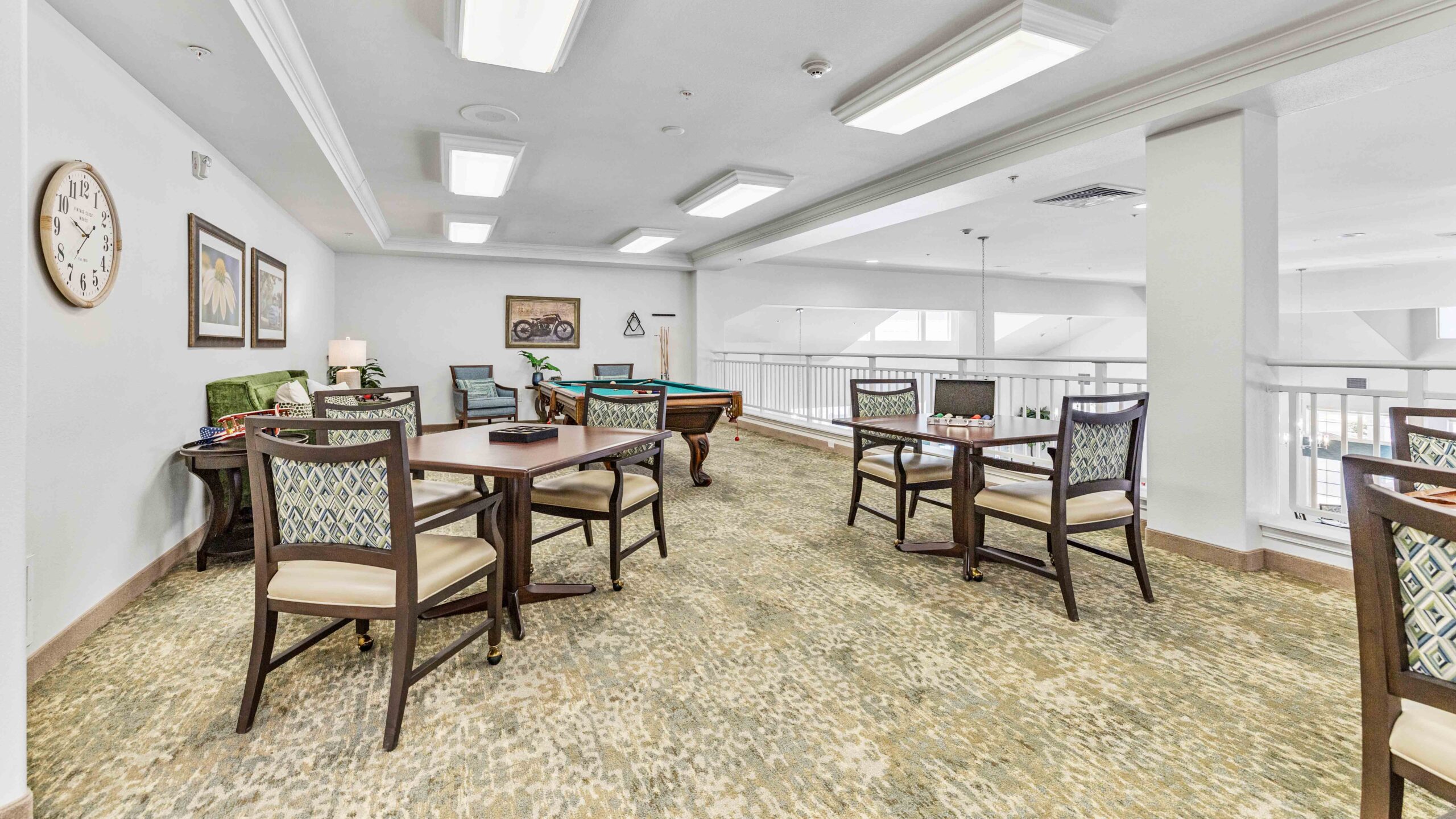 A bright recreation room with tables and chairs, a pool table, chess set, and wall art. Neutral-colored carpet, white walls, and large overhead lights create a welcoming, open atmosphere.