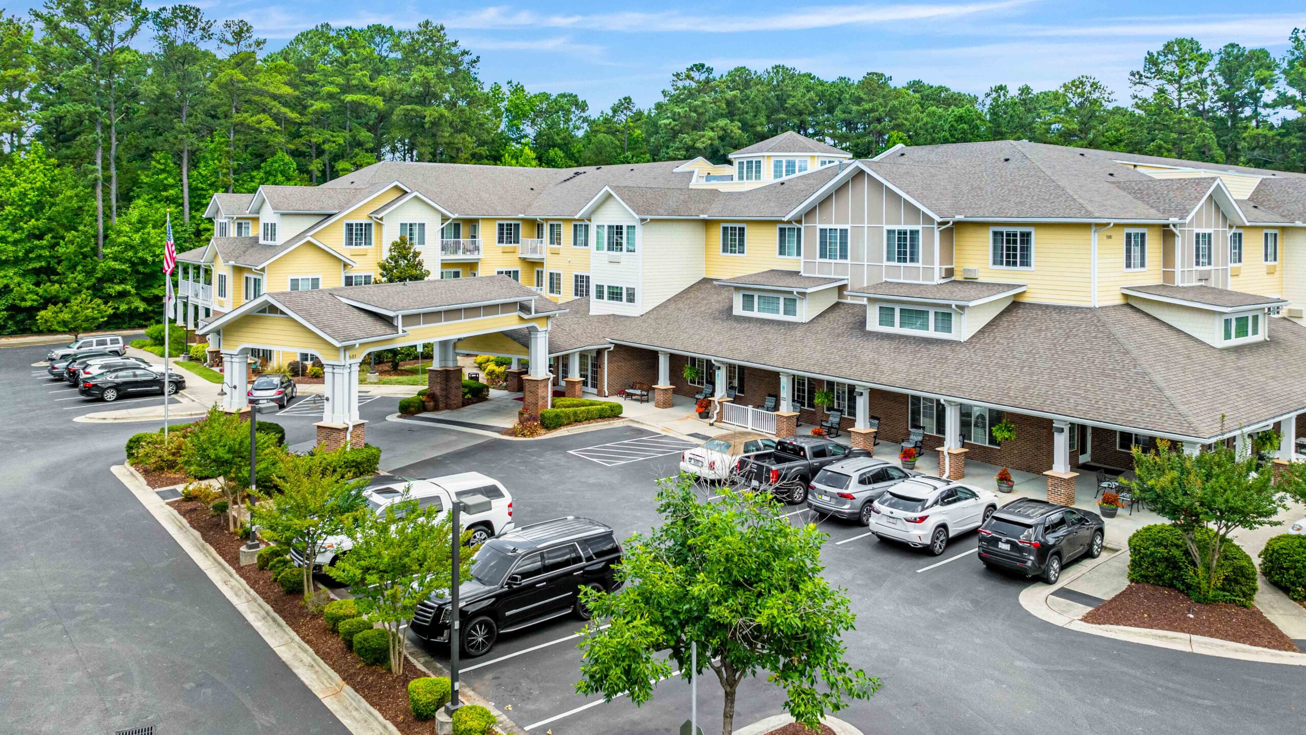 A large, yellow, multi-story building with white trim, likely a residential or assisted living facility, surrounded by trees. Several cars are parked in the lot, and the entrance is covered by a portico.