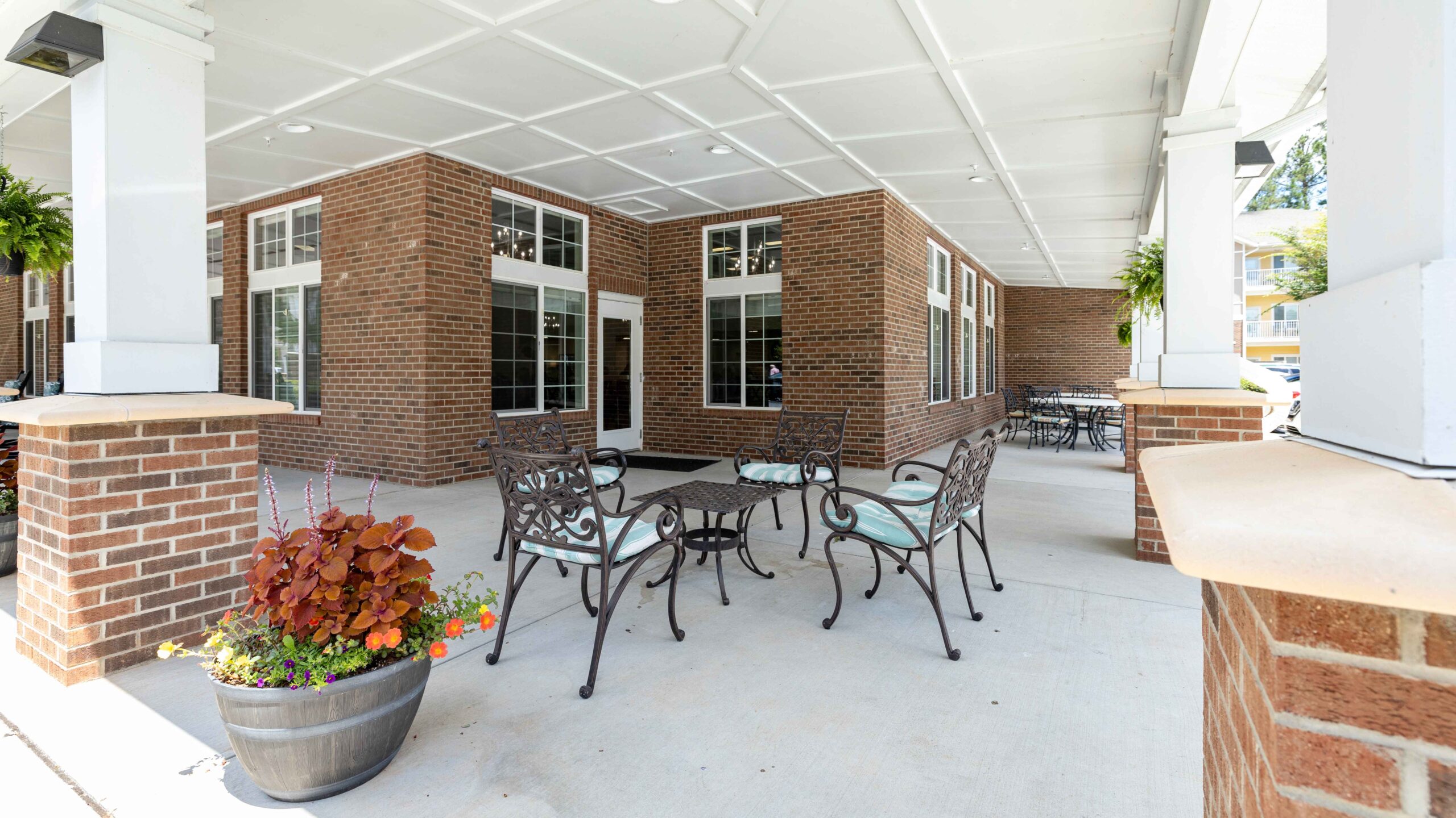 A covered outdoor patio with metal tables and chairs, brick pillars, and large windows. A potted plant with colorful flowers sits in the foreground. The area is bright and inviting.