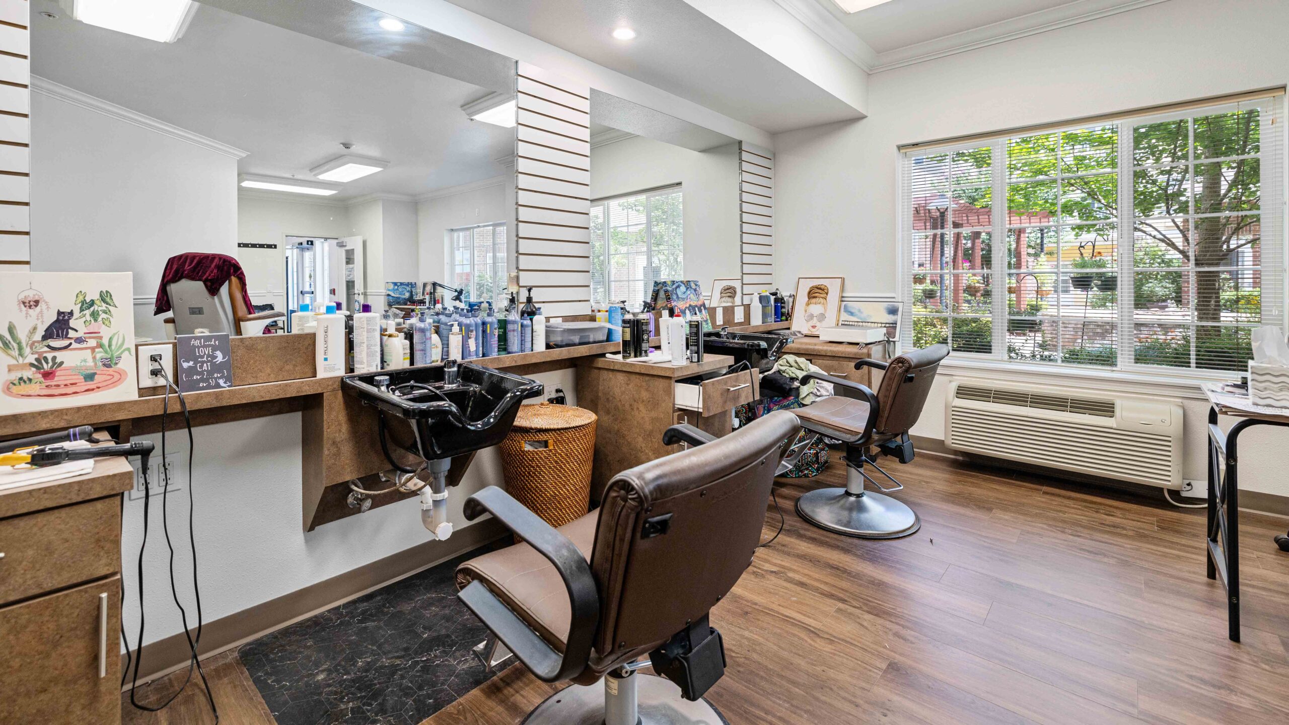 A modern hair salon with two brown styling chairs, mirrors, hair products on countertops, a sink, and large windows letting in natural light, overlooking greenery outside.