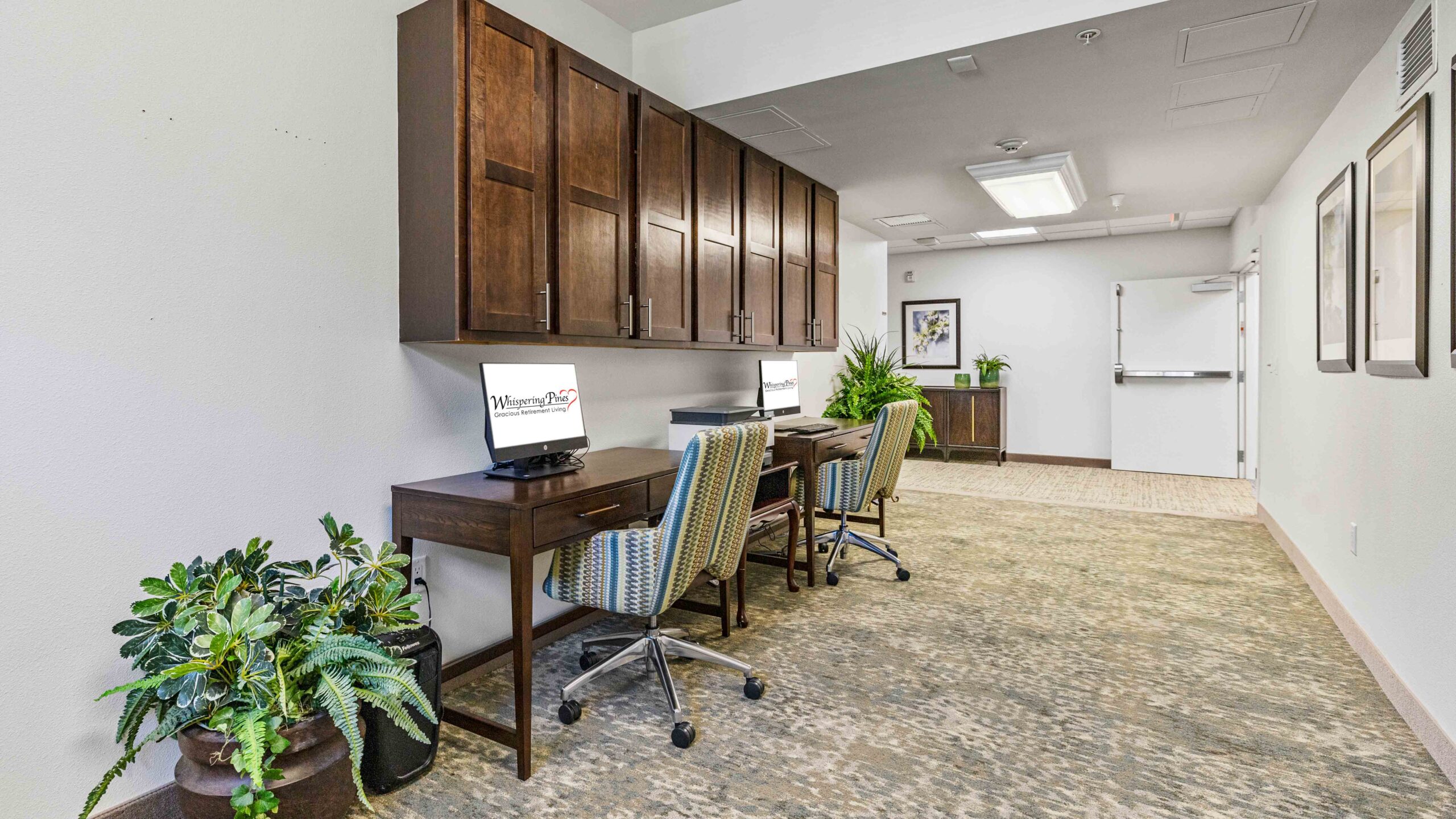 A workspace with two desks and computers, upholstered chairs, potted plants, cabinets above the desks, and art on the walls in a well-lit hallway with carpeted flooring.