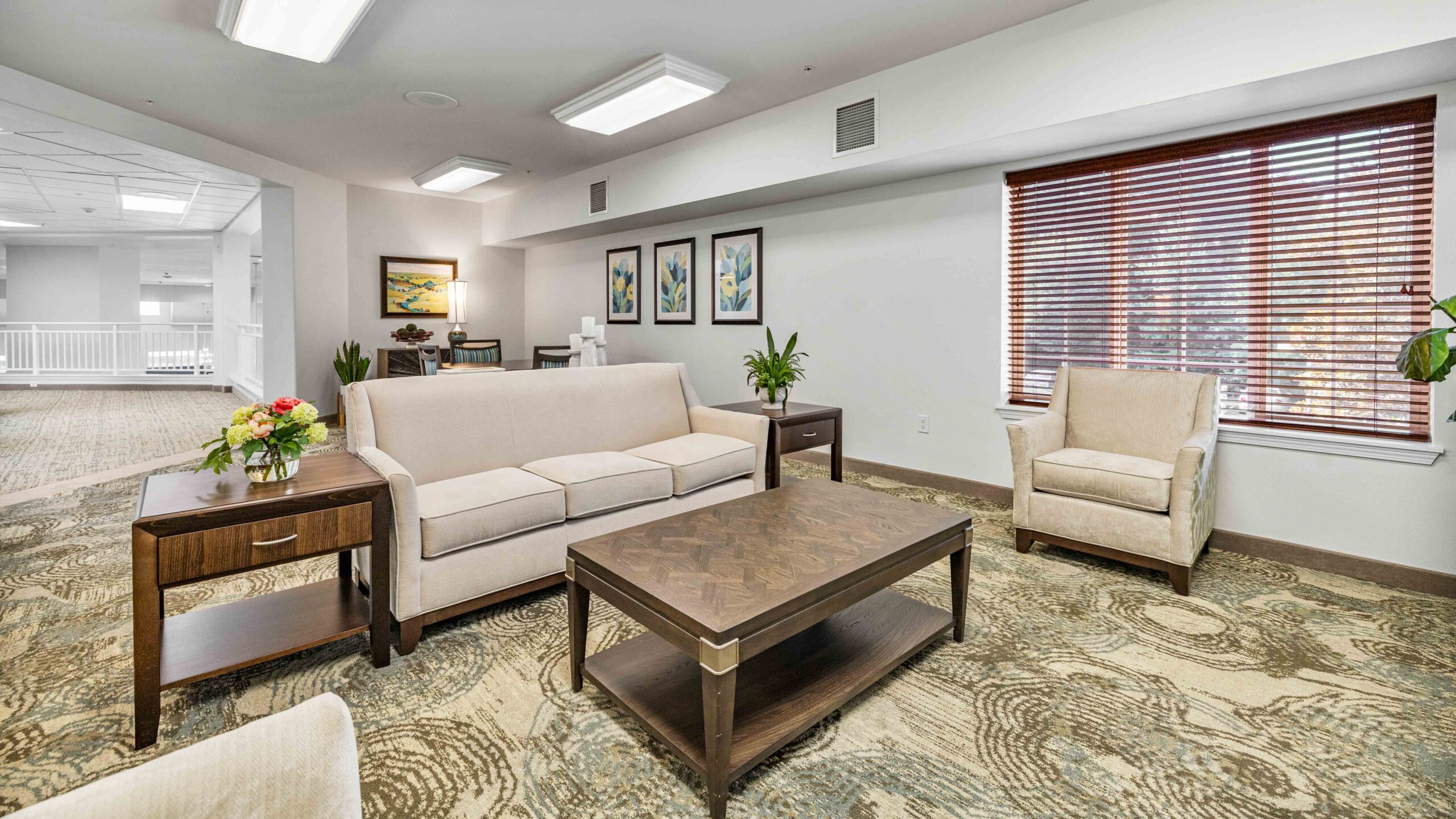 A bright, modern living room with beige sofas, a wooden coffee table, armchair, potted plants, wall art, and a large window with wooden blinds letting in natural light.