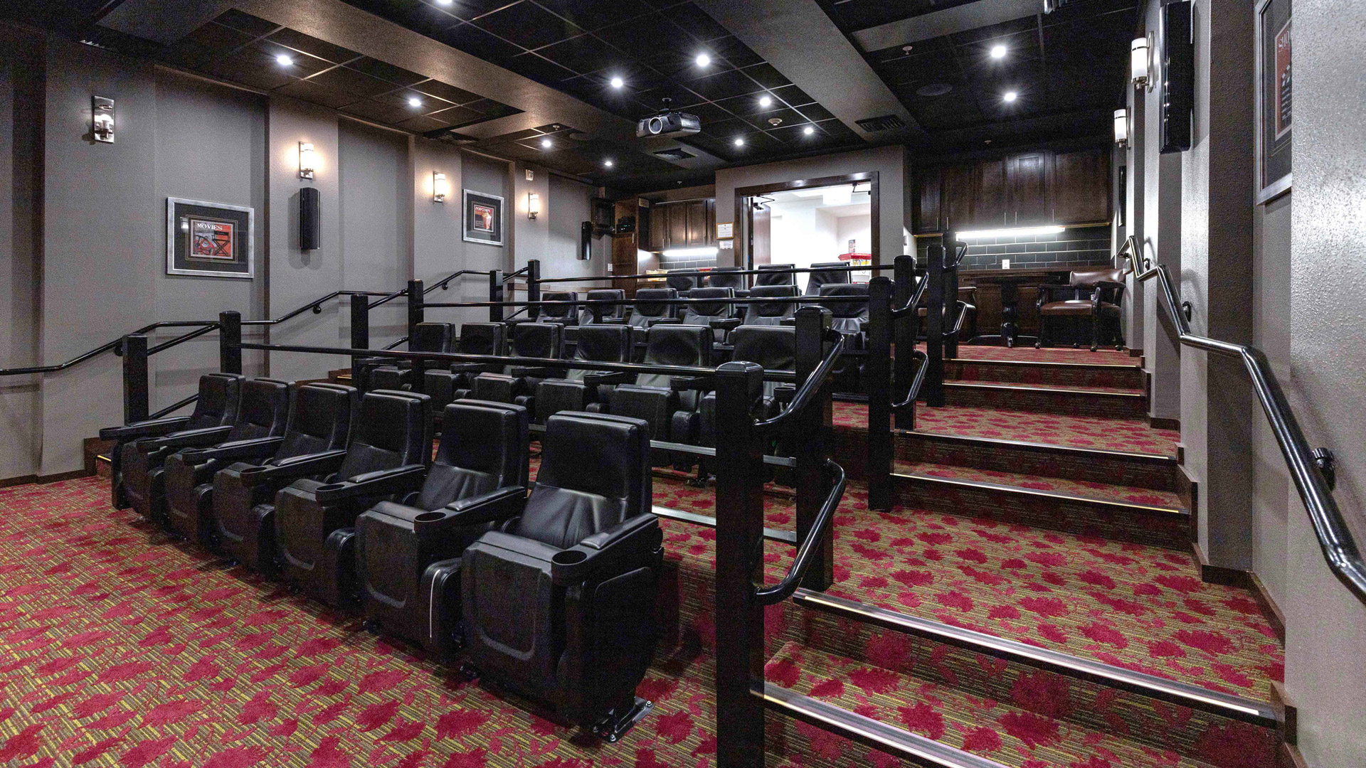 A modern, empty theater with black recliner seats arranged in tiered rows, red-patterned carpet, and dim wall lighting. A projector hangs from the ceiling, and a counter is seen at the back of the room.