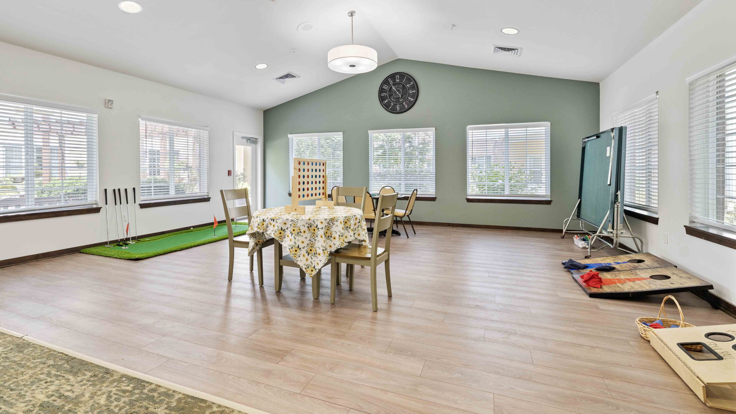 A spacious, bright room with wooden floors, a table covered with a floral cloth, a putting green, a large Connect Four game, and cornhole boards, designed for recreational activities.