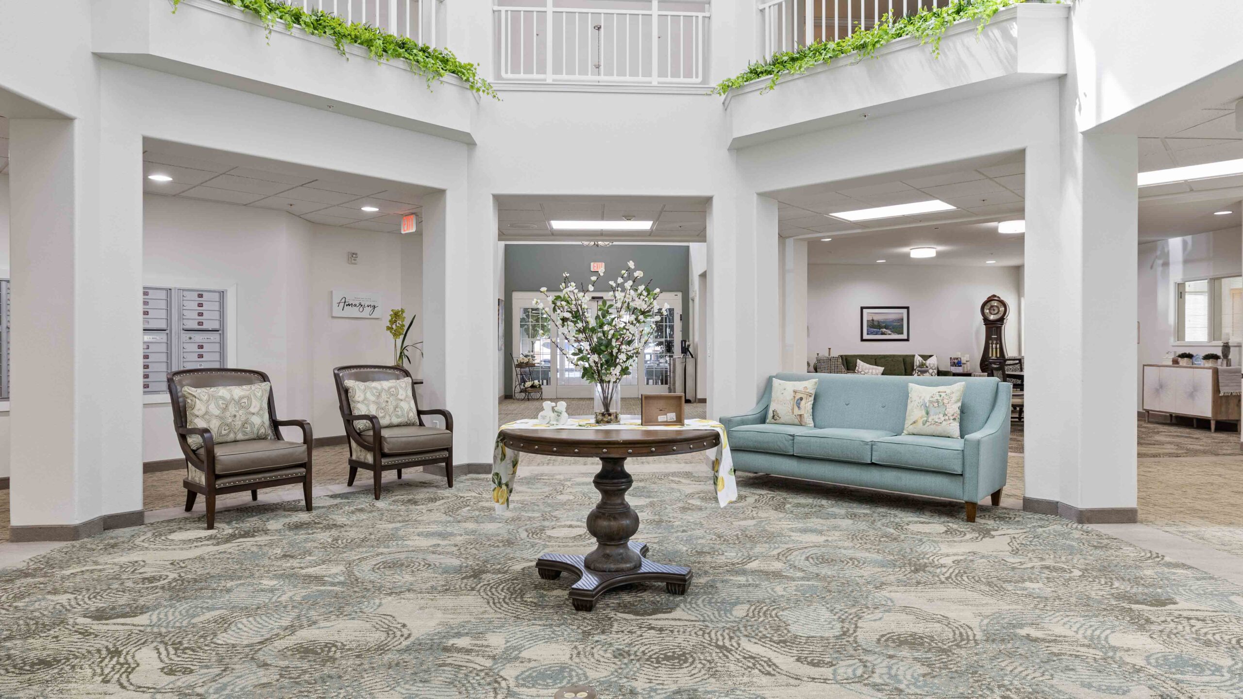 A spacious, well-lit lobby with two armchairs, a round table with flowers, and a blue sofa. Green plants hang from the upper balcony, and patterned carpet covers the floor. The area feels open and welcoming.