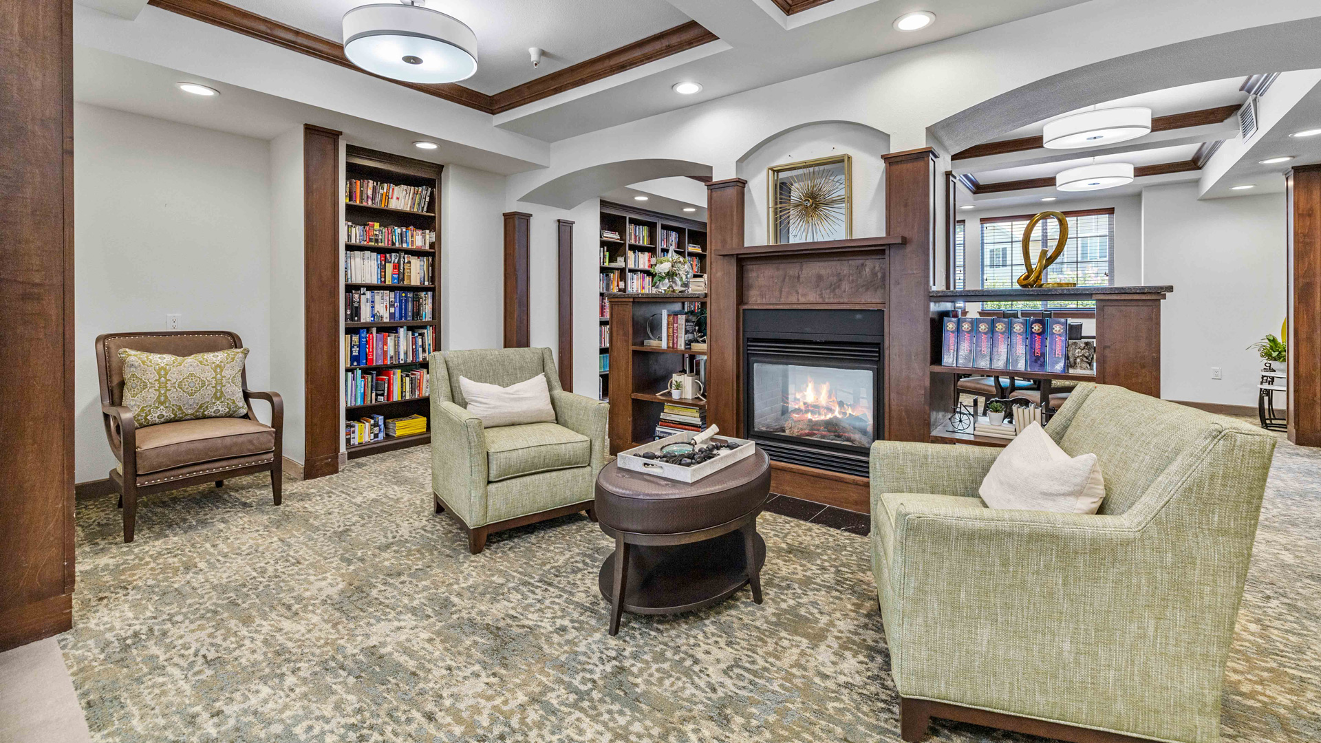 A cozy library with two green armchairs, a round coffee table, a brown armchair, built-in bookshelves, and a double-sided fireplace under a wooden-beamed ceiling with modern light fixtures.