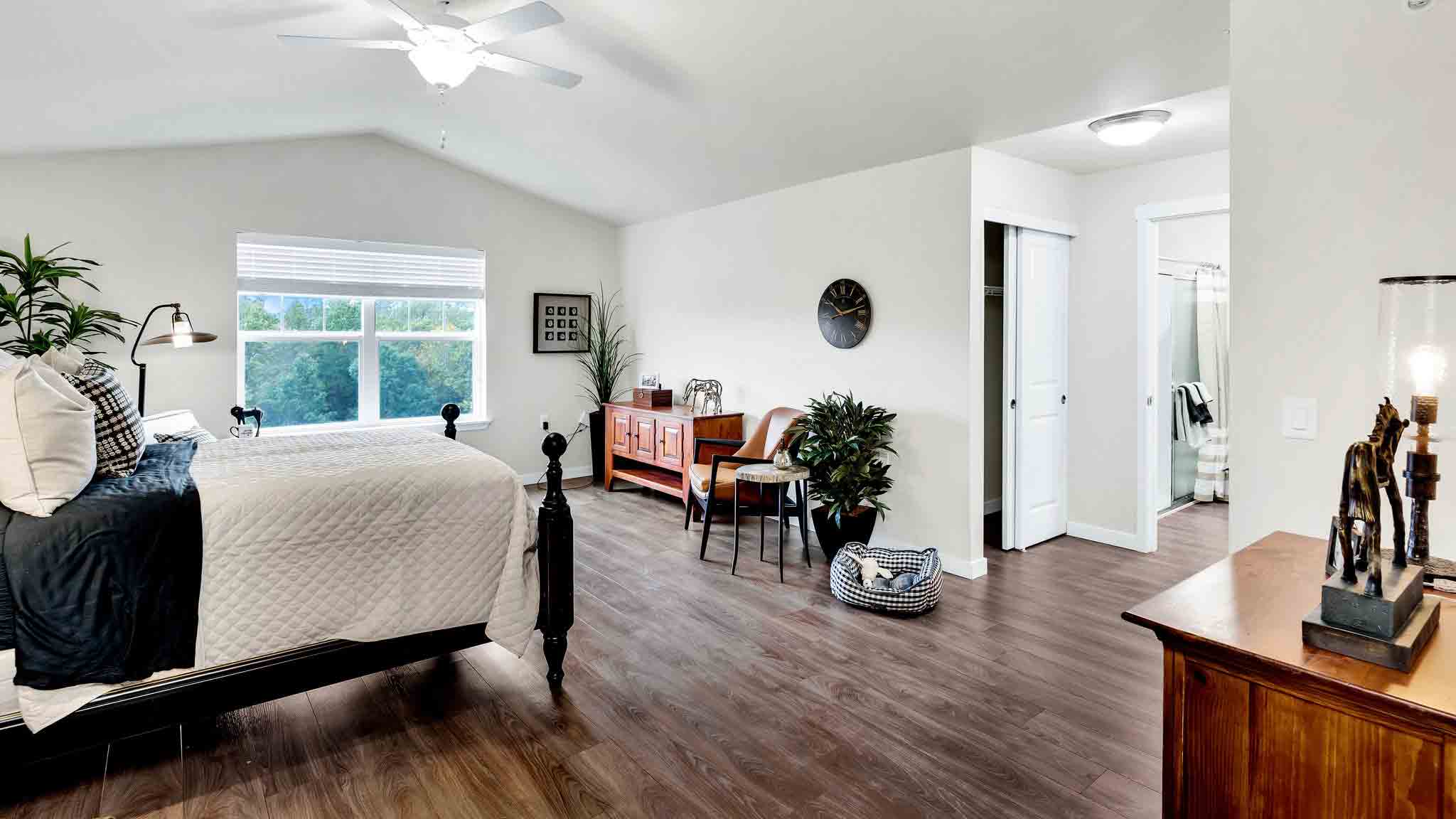 Bright bedroom with a large window, bed with white bedding, wooden floor, dresser, seating area with plants, and an open door leading to a bathroom. A dog bed sits near the seating area.