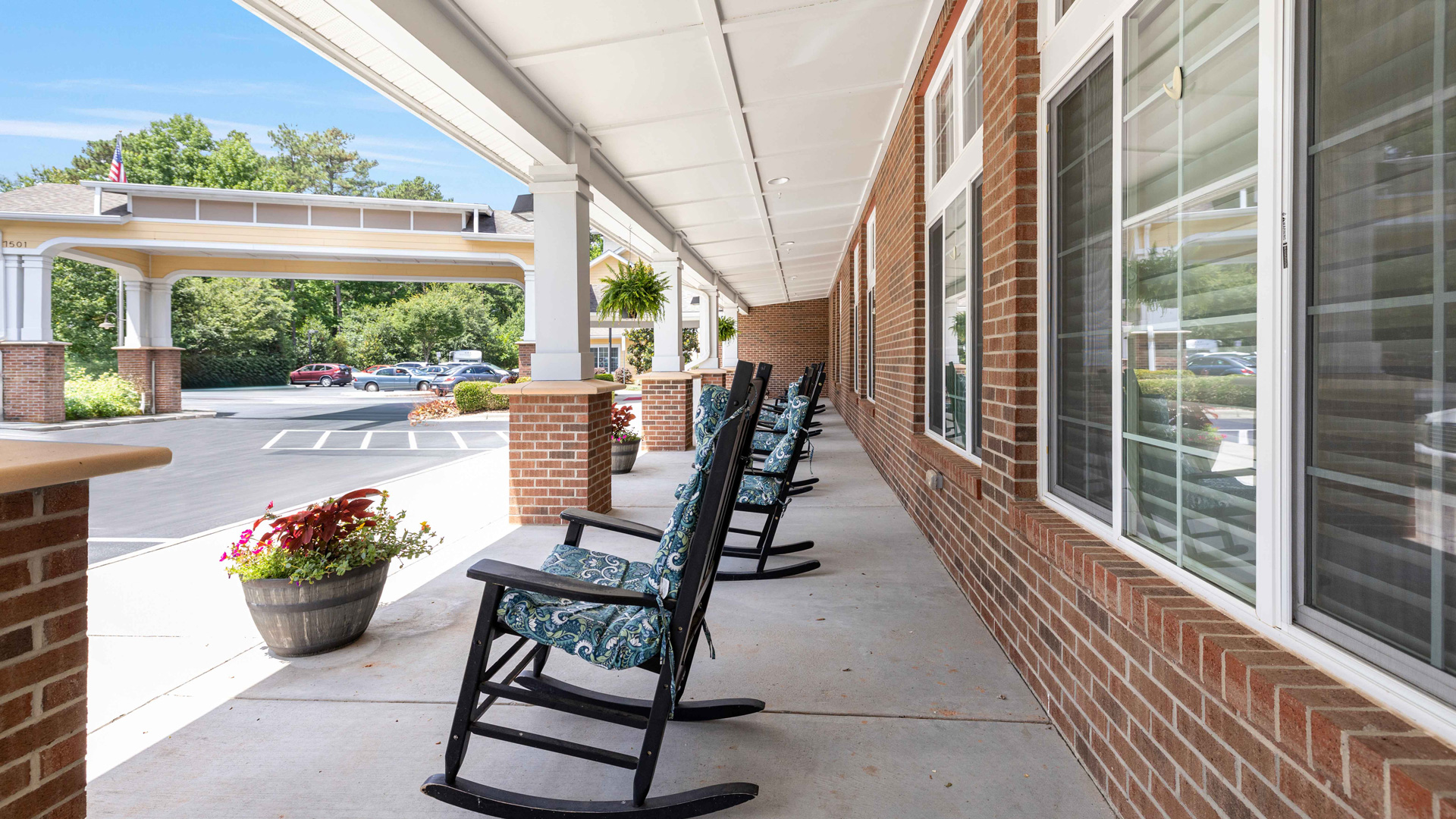 A covered porch with several cushioned rocking chairs lined up along a brick wall, overlooking a parking lot and landscaped greenery on a sunny day.