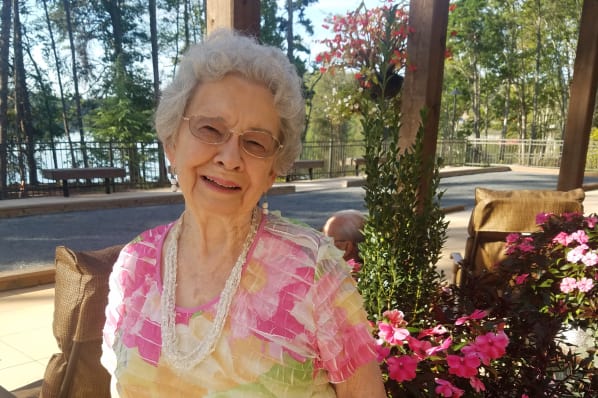 An elderly woman with white hair and glasses is smiling and wearing a colorful floral shirt with a beaded necklace. She is standing outdoors near pink flowers, with trees and a body of water in the background. It is a sunny day.