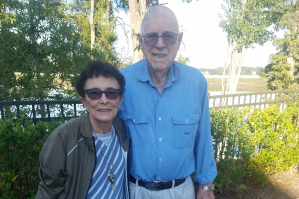 An elderly couple stands outdoors by a railing with a scenic backdrop of trees and water. They both wear eyeglasses; the woman is in a striped shirt with a jacket, and the man is in a blue button-down shirt and light-colored pants. They are smiling at the camera.