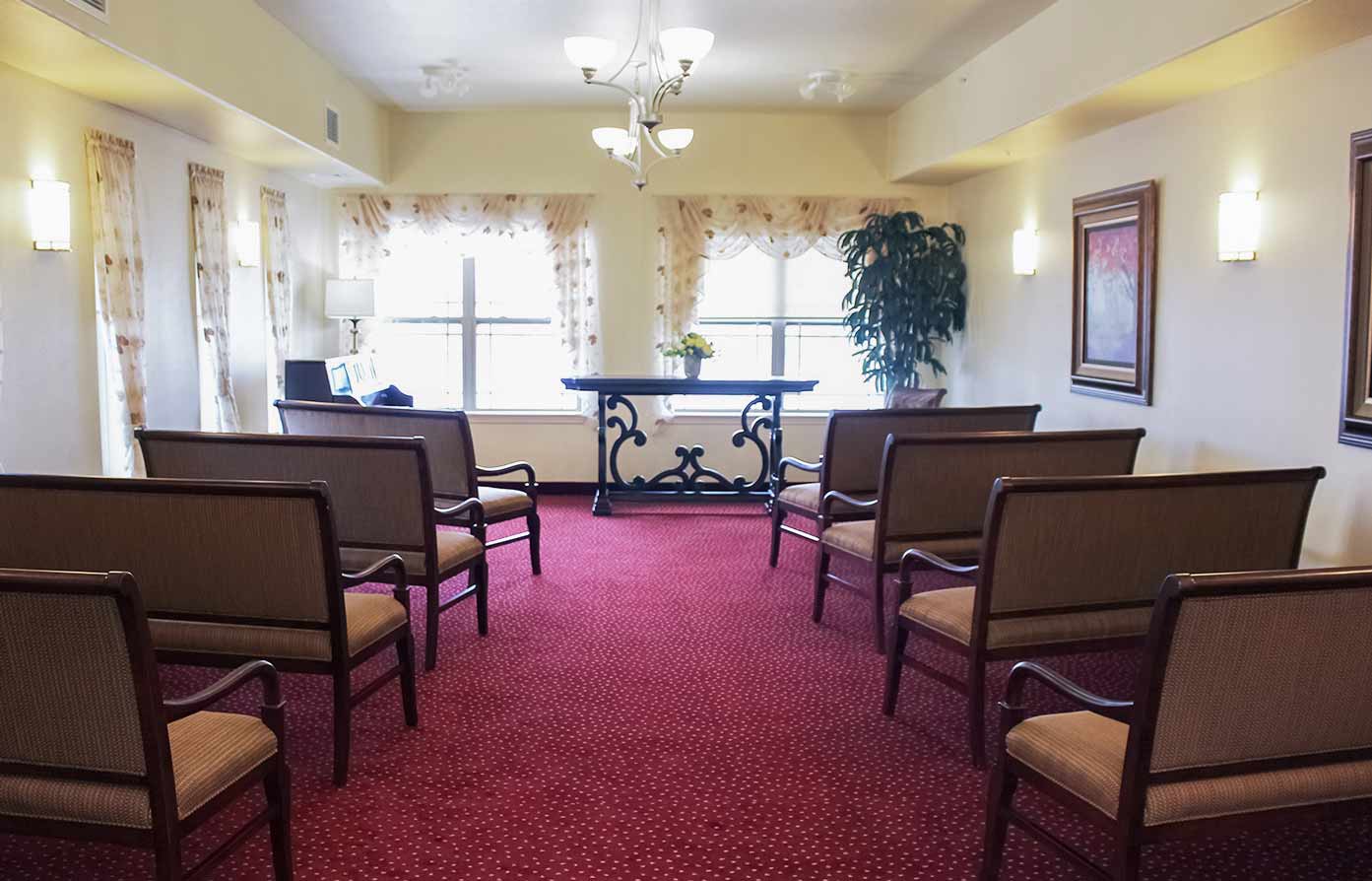 A small chapel with red carpeted floors, several rows of beige cushioned chairs, and an ornate wooden altar at the front. Two large windows with sheer curtains allow natural light to brighten the room. Two framed pictures are on the walls, and a large plant is near the altar.