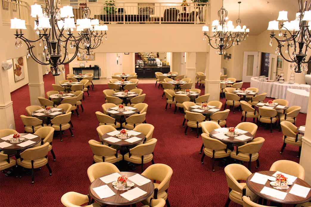 A spacious dining hall with elegant chandeliers, beige upholstered chairs, and round tables set with white tablecloths and red flower centerpieces. The hall features a red carpet and a balcony overlooking the dining area in the background.