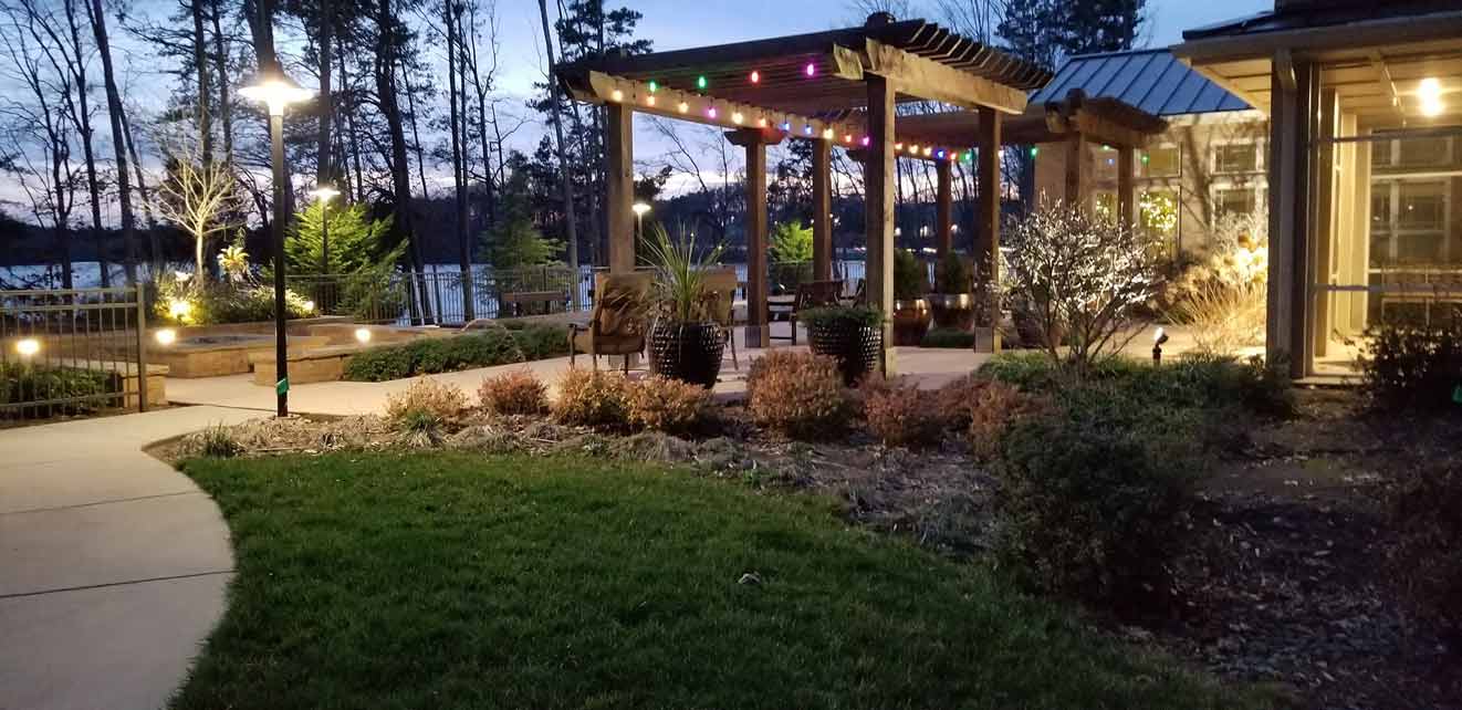 A serene evening scene of a landscaped outdoor area featuring a pergola adorned with colorful string lights, surrounded by various plants and bushes. Pathways and seating areas are illuminated by soft lamplight, with a visible lake and trees in the background.