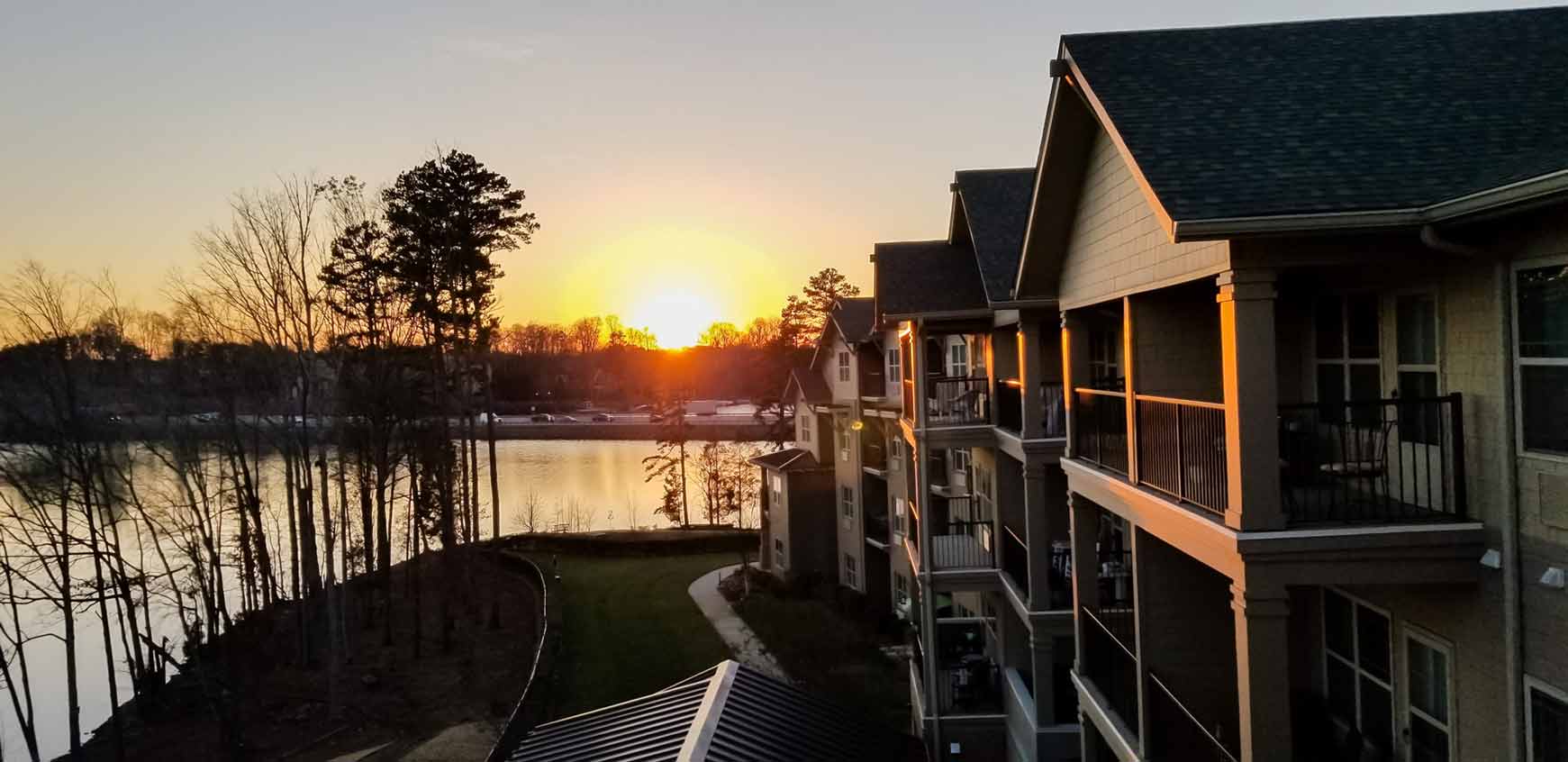 A scenic view of an apartment complex at sunset, showcasing the orange and yellow hues of the sun setting behind trees and a tranquil lake. The buildings cast shadows and reflect the warm sunlight, creating a peaceful atmosphere.