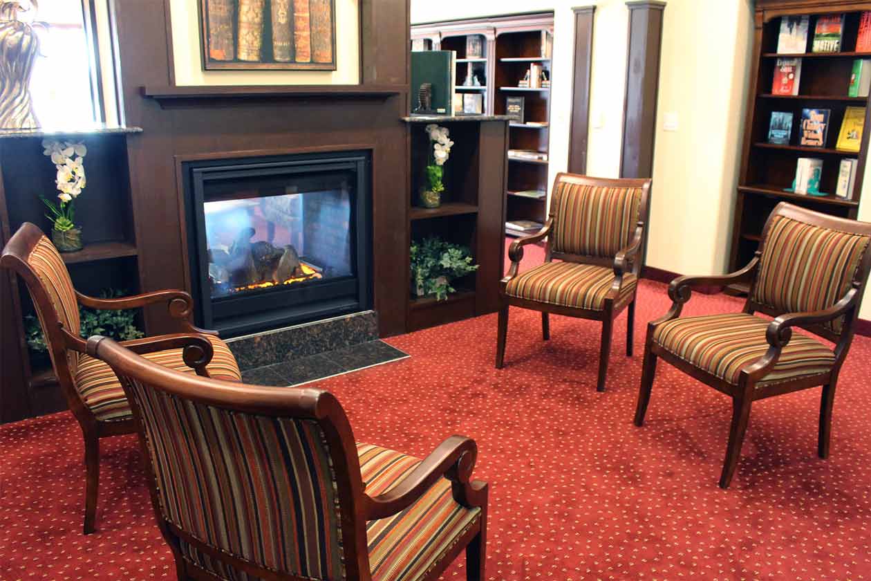 A cozy room features a fireplace surrounded by wooden bookshelves filled with various books. Four striped, upholstered armchairs are arranged in a semi-circle on a red, speckled carpet, creating a comfortable reading or conversation area.