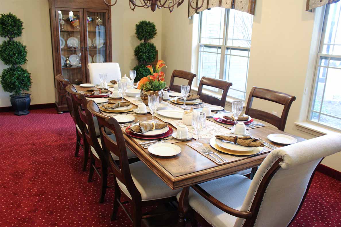 A dining room with a wooden table set for ten, featuring beige cushioned chairs, polished silverware, and folded napkins. A floral centerpiece adorns the table. A hutch with glass doors and decor items is in the corner, with potted plants adding greenery to the space.