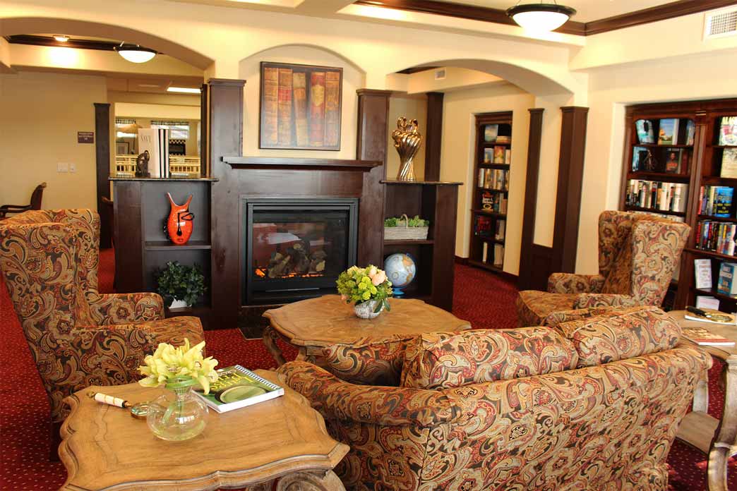 A cozy, well-lit library with red carpet features a lit fireplace surrounded by bookshelves. There are two patterned armchairs and a matching sofa around wooden coffee tables, adorned with decorative flowers, a globe, and books.