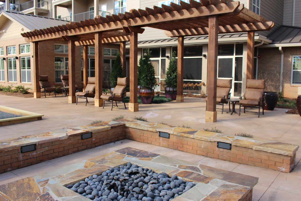 Outdoor patio with wooden pergola, lounge chairs, and a brick rectangular fire pit filled with smooth black stones. Tall plants in pots are placed against the building, and the backdrop features large windows and a garden area.