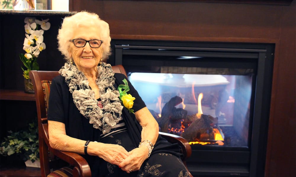 An elderly woman with white hair and glasses sits smiling in a chair next to a fireplace. She is wearing a black dress, a lace scarf, and a yellow corsage. The background includes a bookshelf with plants and decor.