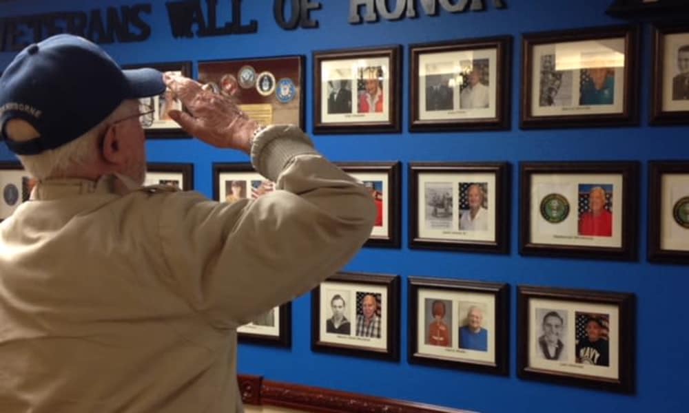 An elderly man in a blue cap salutes a wall displaying framed photos of veterans, labeled 