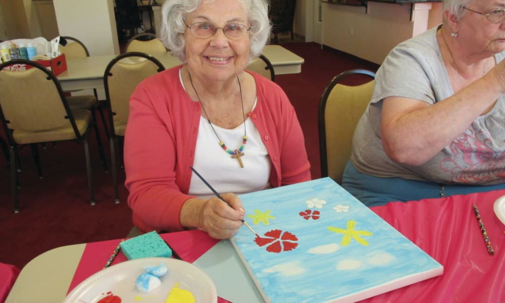 An elderly woman with white hair and glasses, wearing a bright pink cardigan, is smiling and painting a canvas with flowers and star shapes in a room with tables and other people. She holds a paintbrush, and the table is covered with art supplies.