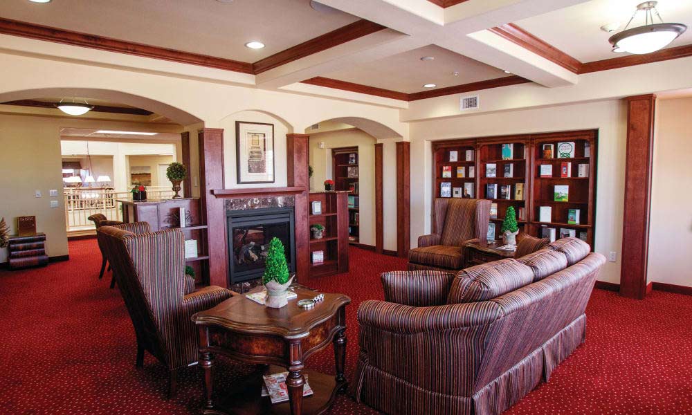 A cozy living room featuring a red carpet, striped sofas, armchairs, and a wooden coffee table. Bookshelves filled with books line the walls. A fireplace with a decorative mantle is central, with potted plants and framed artwork enhancing the elegant ambiance.