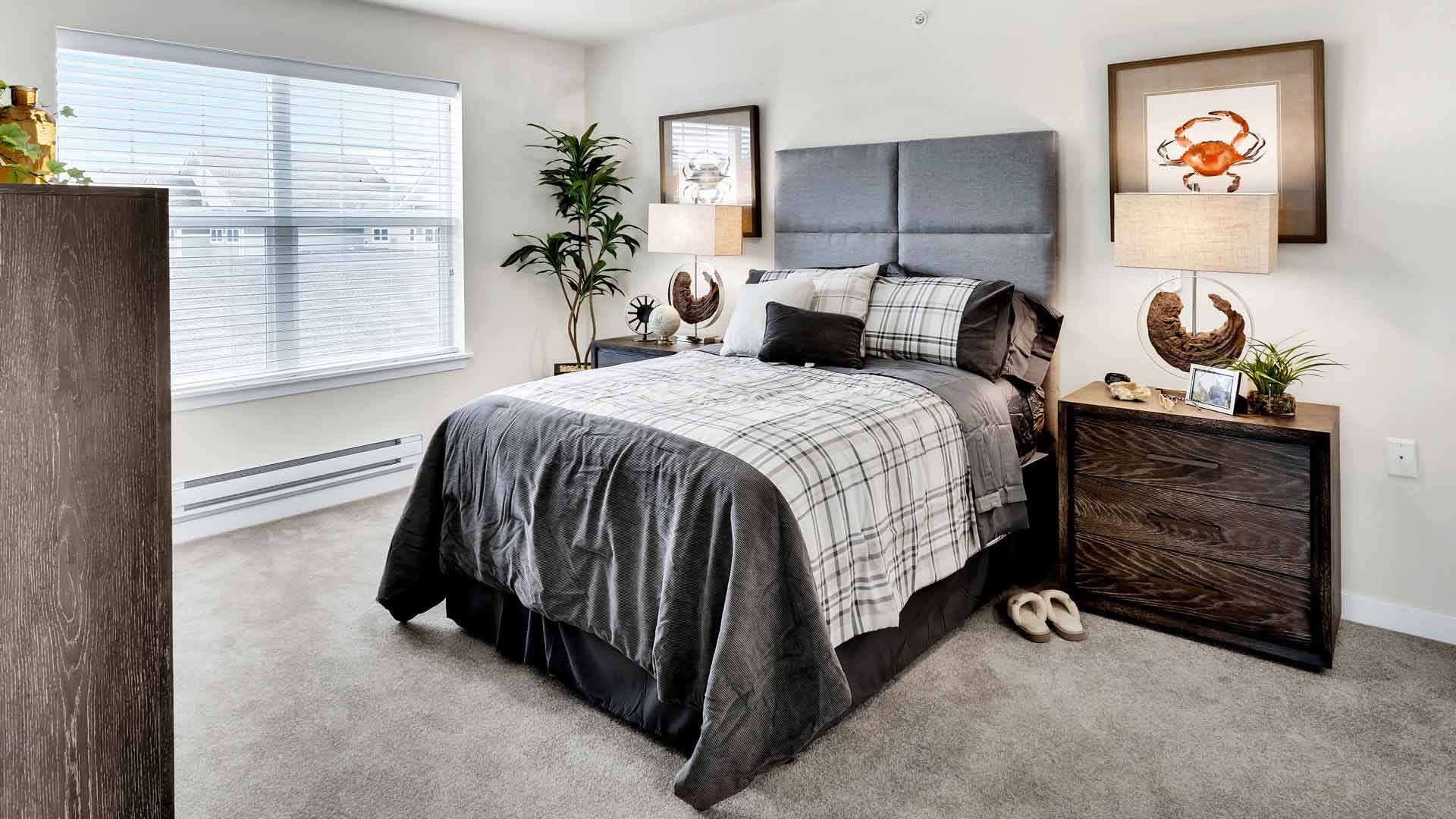 A modern bedroom with a double bed covered in gray and plaid bedding. Two wooden nightstands with lamps flank the bed. Artwork with crab designs hangs above them. A large window on the left lets in natural light. A plant is in the corner.