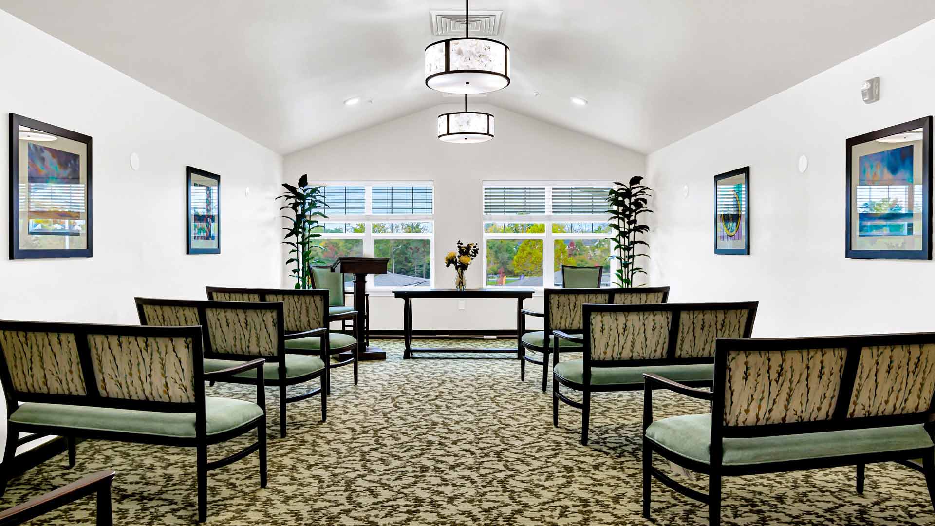 A small, bright chapel with a vaulted ceiling features six rows of dark wooden chairs with light green cushions. Framed artwork adorns the white walls, and two potted plants flank a podium at the front. Three windows let in natural light.