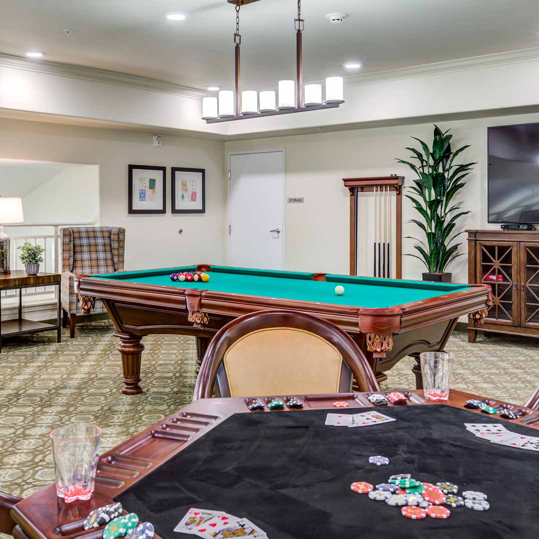 A recreation room featuring a pool table with balls arranged on it and a poker table with playing cards and drinks. The room has a large plant, a wall-mounted TV, a plaid armchair, framed art, and a modern light fixture.