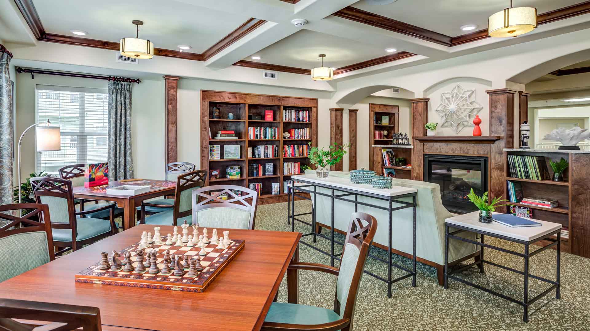 A cozy community room featuring wooden tables with chess sets, bookshelves lined with colorful books, green upholstered chairs, and a fireplace with decorative accents. Warm lighting and large windows add to the inviting atmosphere.