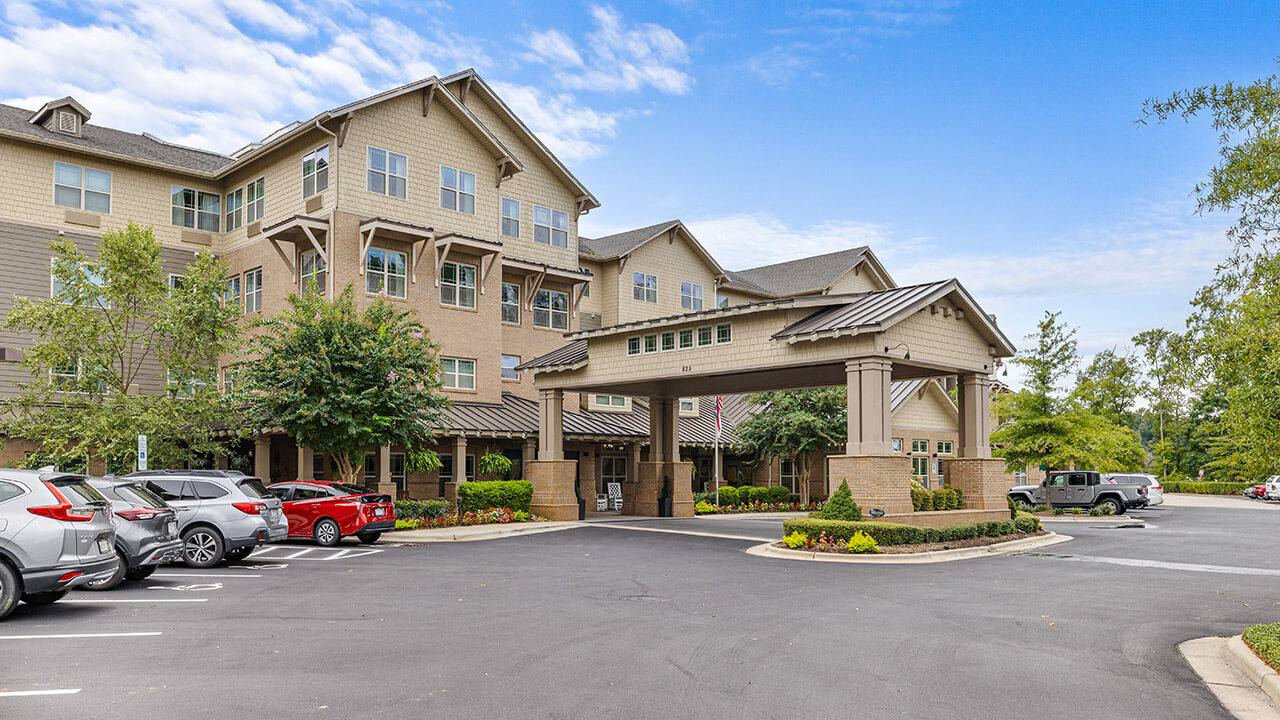 A multi-story beige building with balconies and a covered entrance, surrounded by trees, neatly trimmed bushes, and a parking lot with several parked cars on a bright, partly cloudy day.