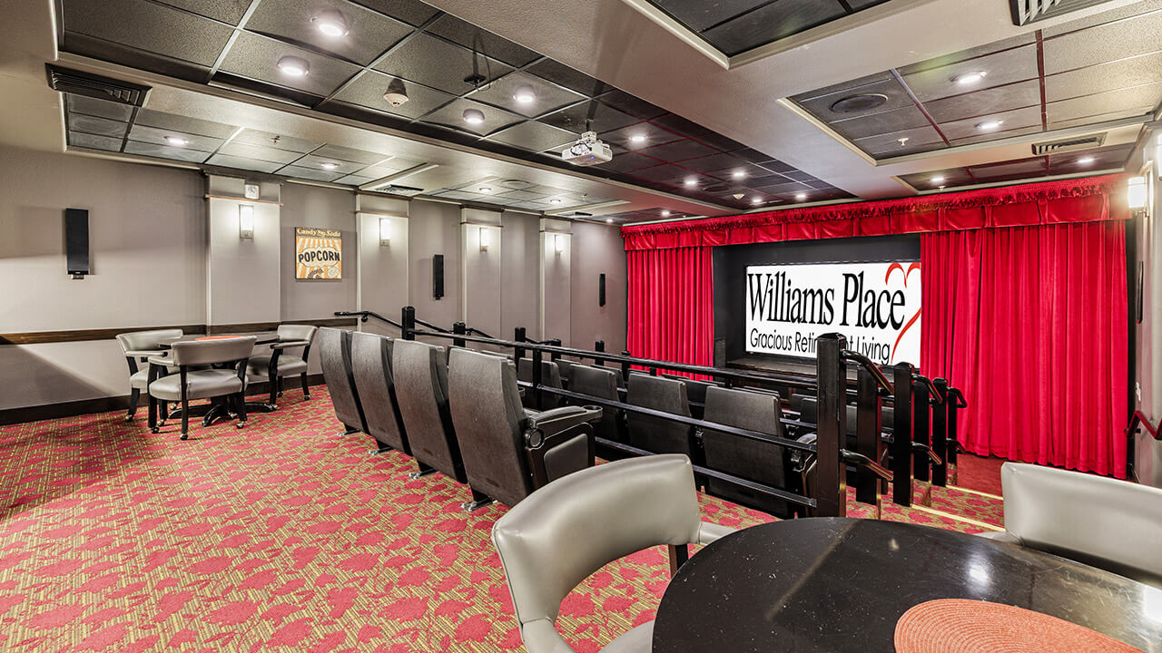 A small movie theater with rows of black recliner chairs, red carpet, round tables with chairs in the foreground, and a screen displaying "Williams Place" with red curtains on the sides.