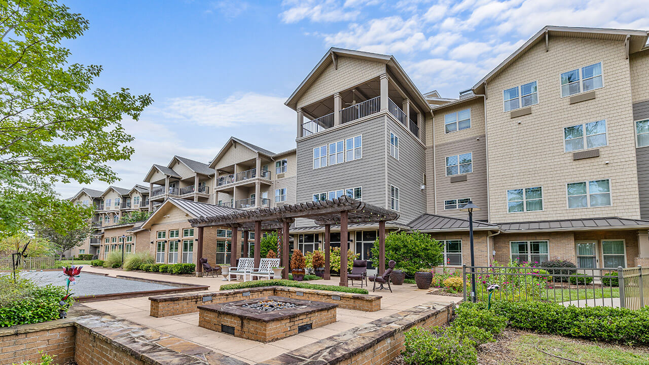 A large, modern apartment building with multiple floors, balconies, and beige siding, surrounded by green landscaping. In the foreground, there is a patio area with seating, a fire pit, and a wooden pergola.