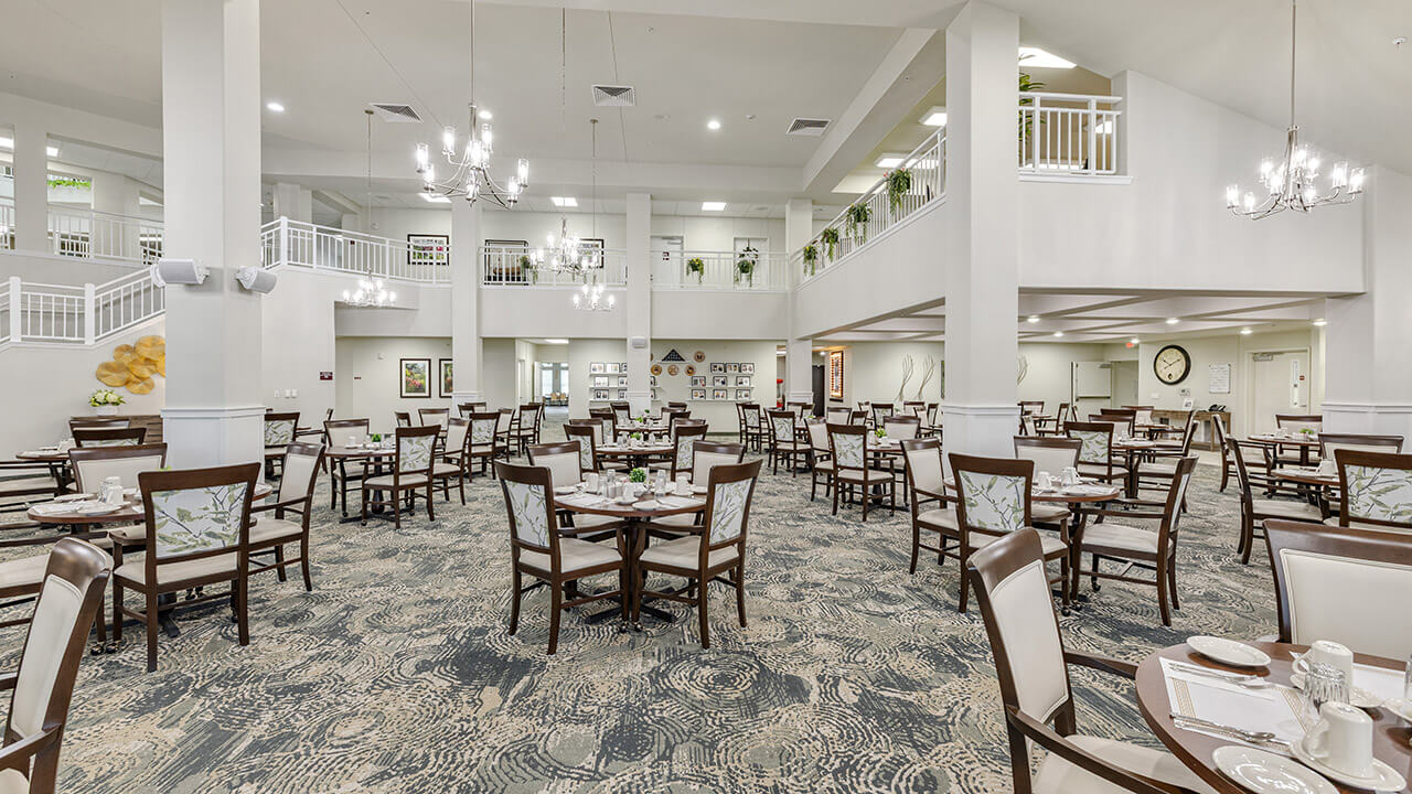 Spacious, well-lit dining area with multiple tables and chairs, elegant chandeliers, white walls, large windows, and a second-floor balcony. The setting is clean, modern, and designed for communal dining.