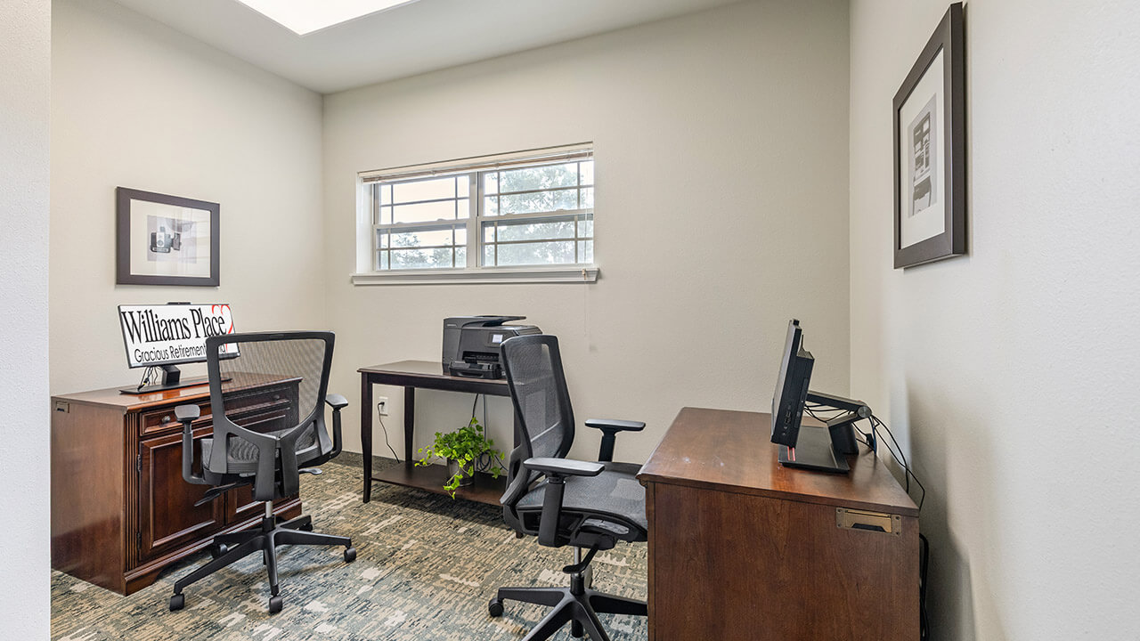 A small office with two wooden desks, two black office chairs, a printer, a computer, framed pictures on the walls, a small plant on the floor, and a window letting in natural light.