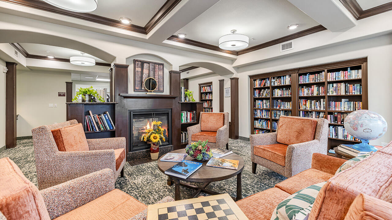 Cozy library lounge with four orange armchairs around a coffee table, a lit fireplace, bookshelves filled with books, a chessboard, magazines, plants, and a globe on a side table. Warm, inviting atmosphere.