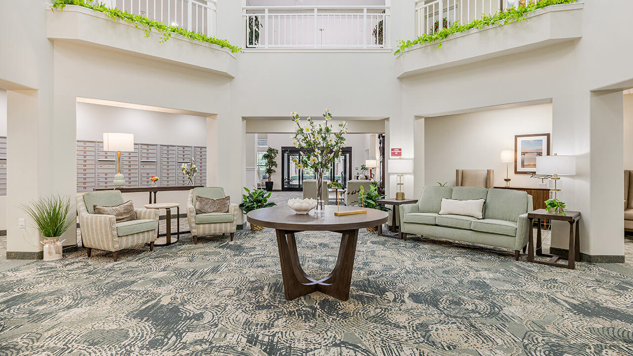A spacious, well-lit lobby with patterned carpet, green sofas, armchairs, plants, a round wooden table with decor in the center, and bright walls with balcony railings and greenery above.