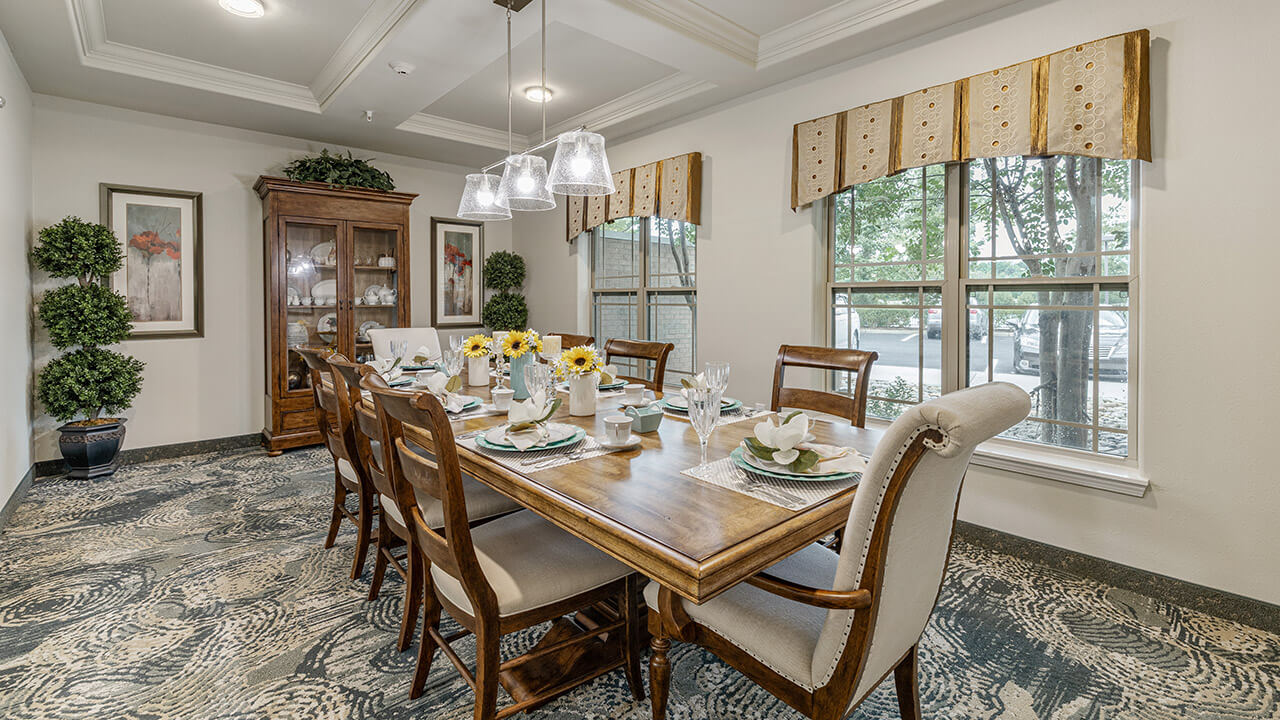 A bright dining room with a long wooden table set for eight, surrounded by chairs. Large windows let in natural light. There are decorative plants, a china cabinet, and floral accents creating a welcoming atmosphere.