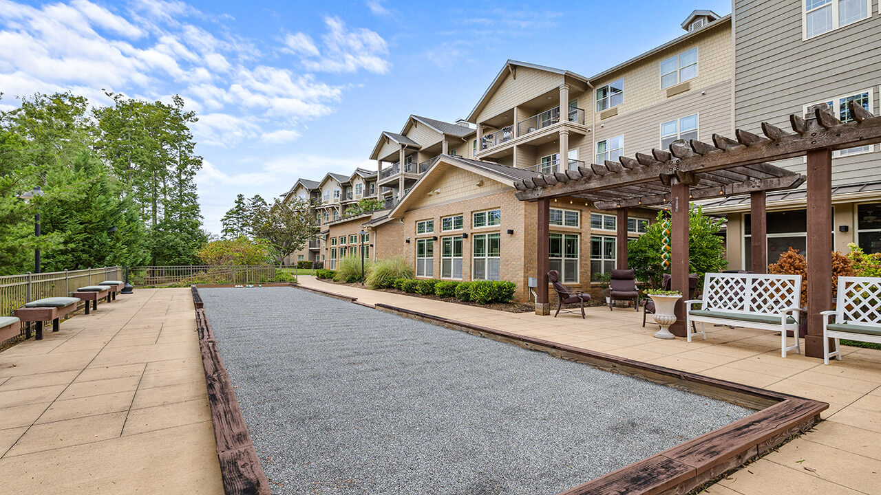 A bocce ball court runs alongside a patio with benches and a pergola outside a multi-story residential building, surrounded by greenery under a partly cloudy sky.