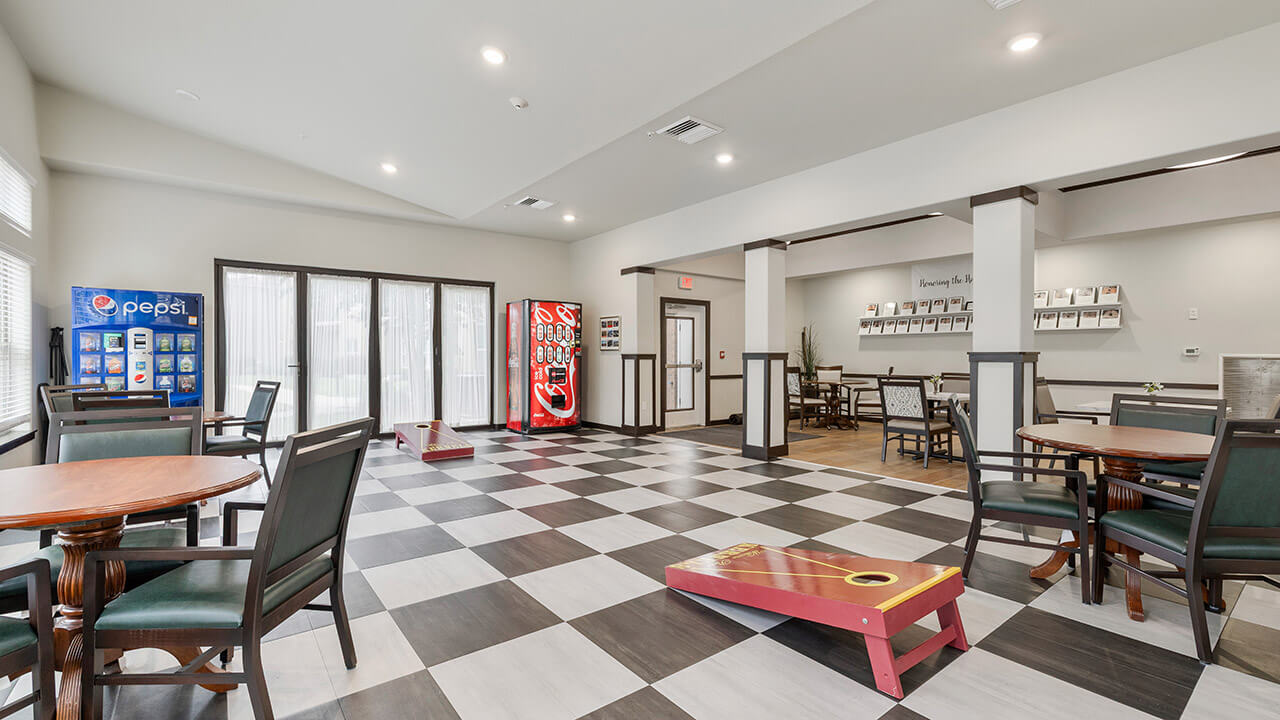 A spacious room with black-and-white checkered flooring, round tables with chairs, a Pepsi vending machine, a Coca-Cola machine, a cornhole game board, and a dining area in the background.
