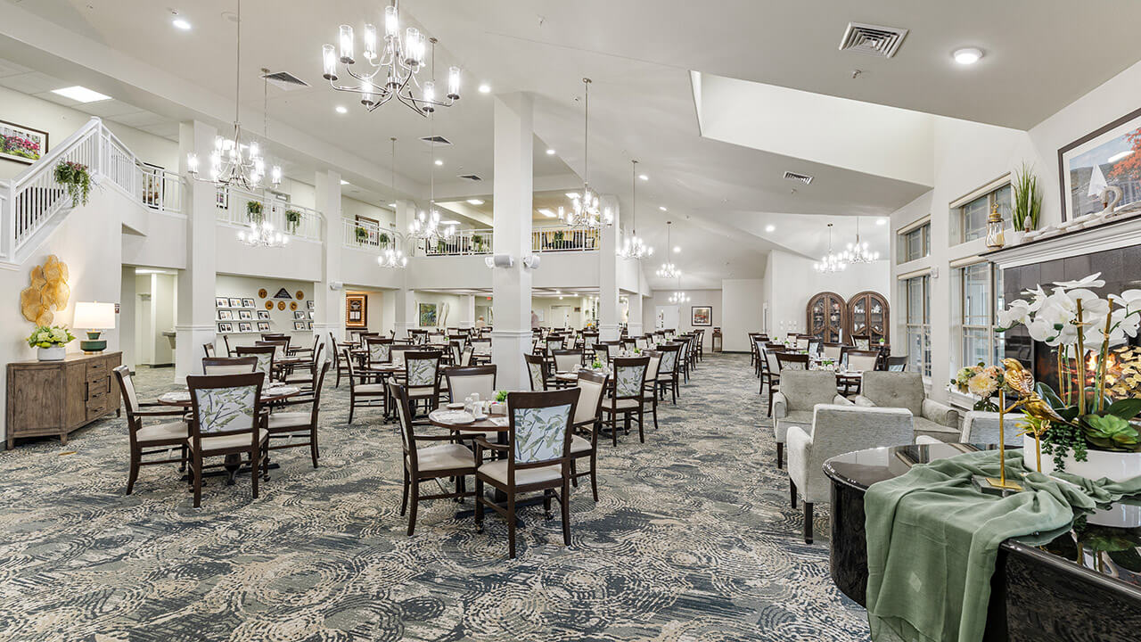 A spacious, elegant dining room with high ceilings, chandeliers, multiple tables and chairs, large windows, and tasteful decor. The room is bright, airy, and set up for communal dining.