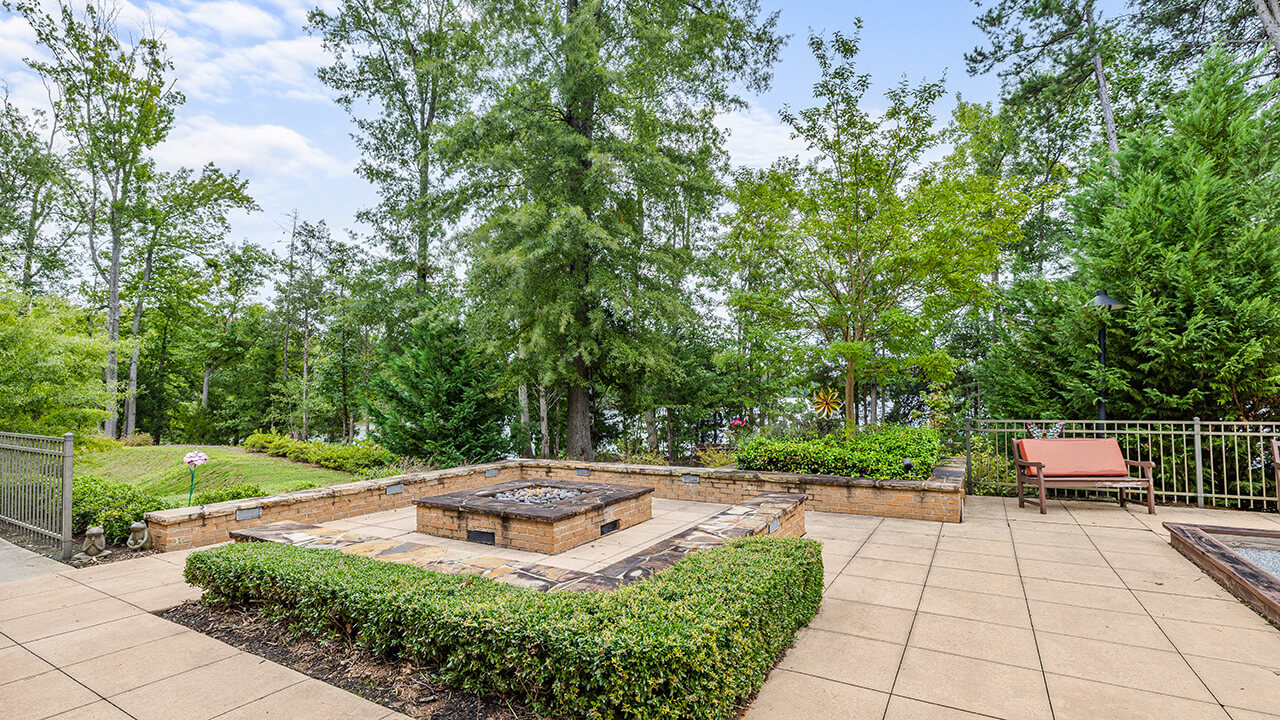 A paved patio features a square fire pit bordered by low hedges and stone seating walls, surrounded by lush green trees and a metal fence, with a brown outdoor lounge chair on the right.