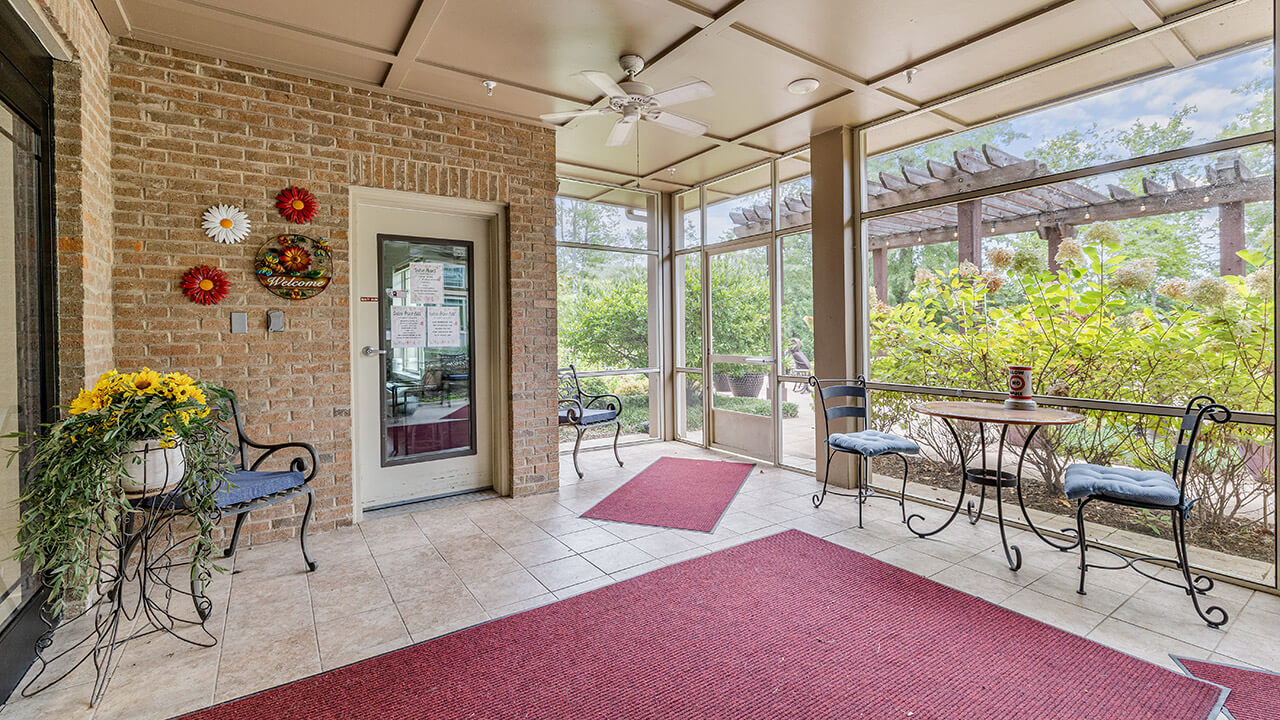 A sunroom with tiled floors, red rugs, brick walls, potted flowers, and black metal chairs with blue cushions. Large windows and a glass door provide views of greenery and a pergola outside.
