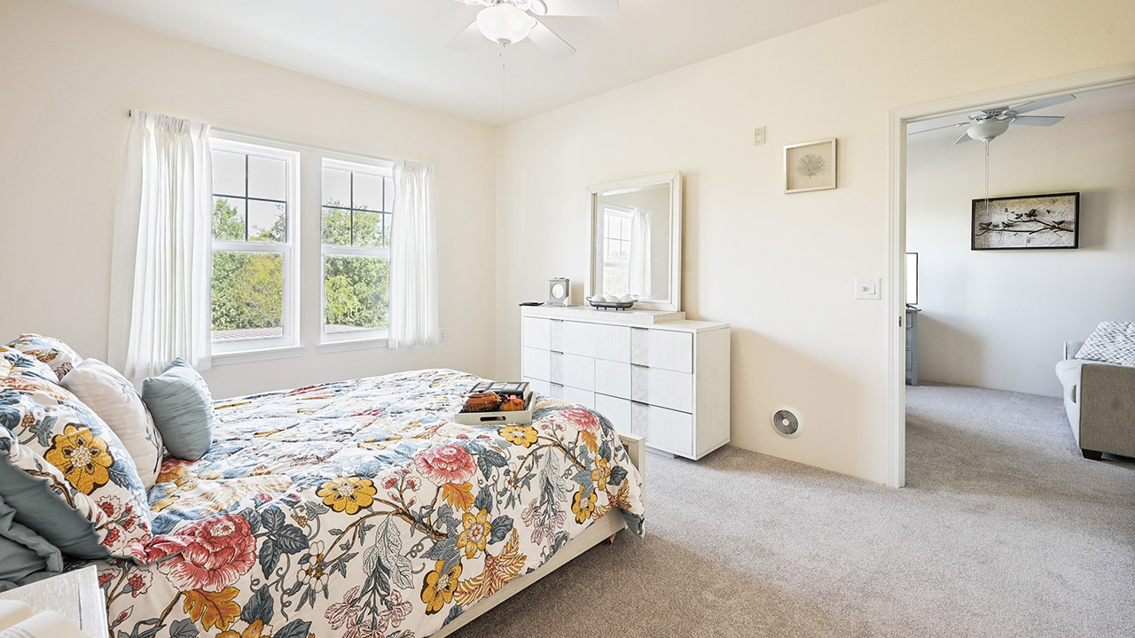 Bright bedroom with a floral-patterned bedspread, white dresser with a mirror, large window with white curtains, and beige carpet. An open doorway leads to another room with a gray sofa.