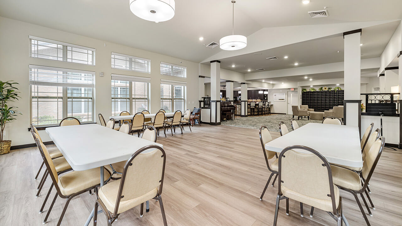 A spacious, well-lit dining room with several rectangular tables and beige chairs on a light wood floor. Large windows let in natural light, and modern ceiling lights hang above the open, airy space.