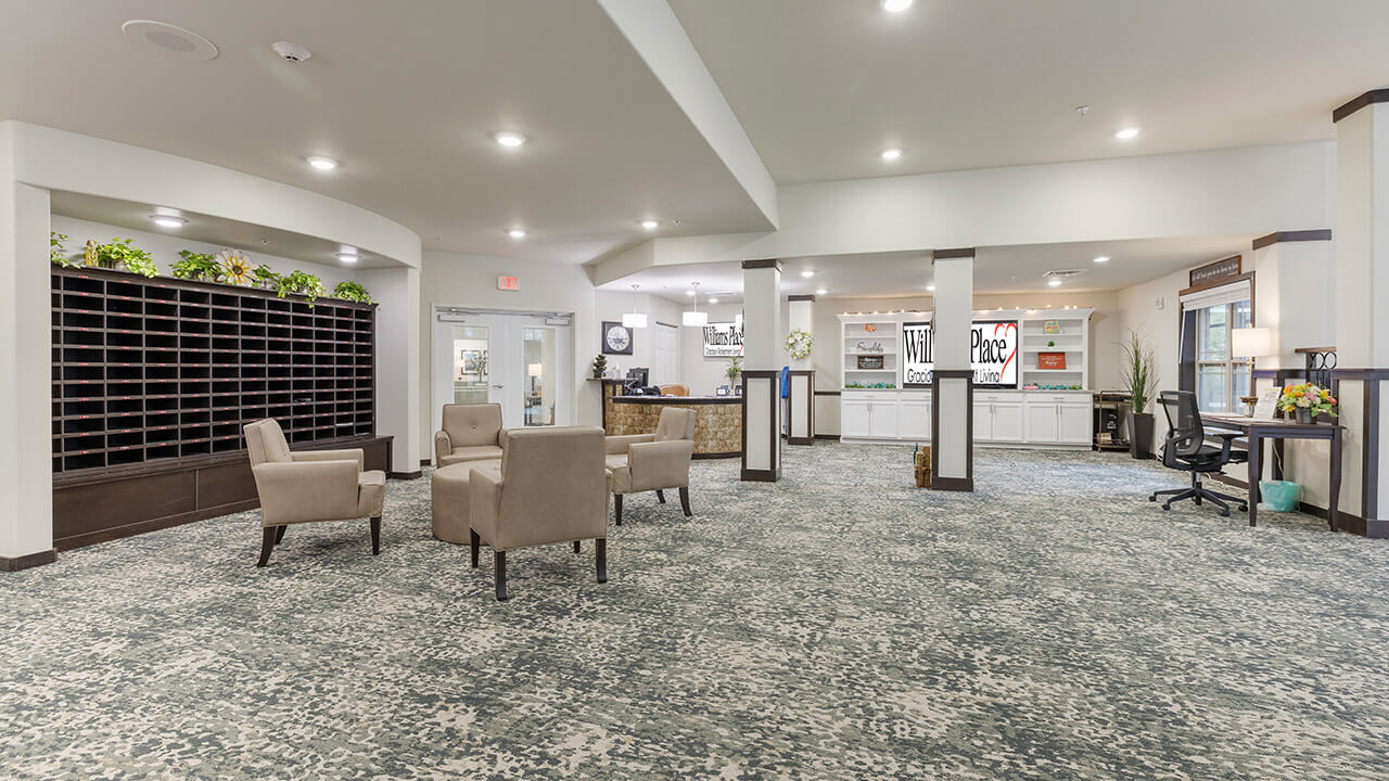 A spacious, well-lit lobby with patterned carpet, beige chairs arranged in small groups, a reception desk, wall-mounted mailboxes, and decorative plants on high shelves.