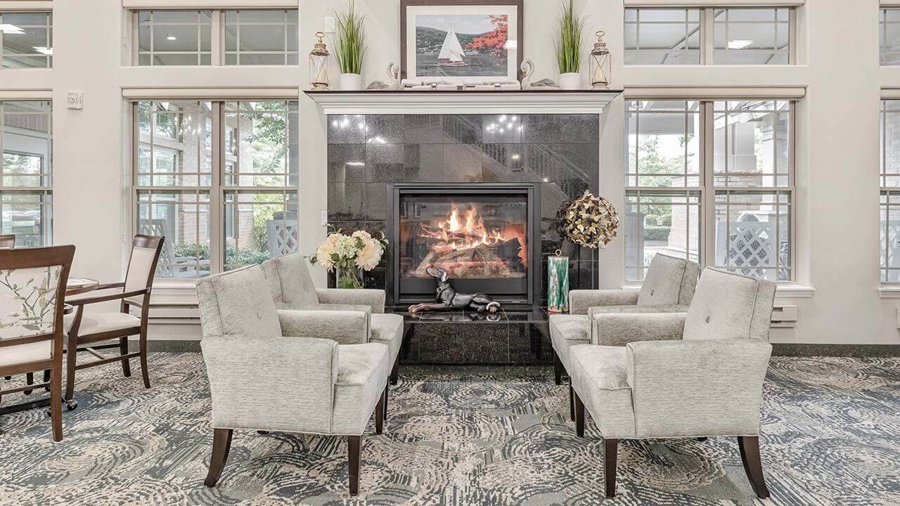 A cozy lounge with four light gray armchairs arranged around a black fireplace. Flowers and decor sit on the mantel, and large windows let in natural light, creating an inviting, elegant atmosphere.