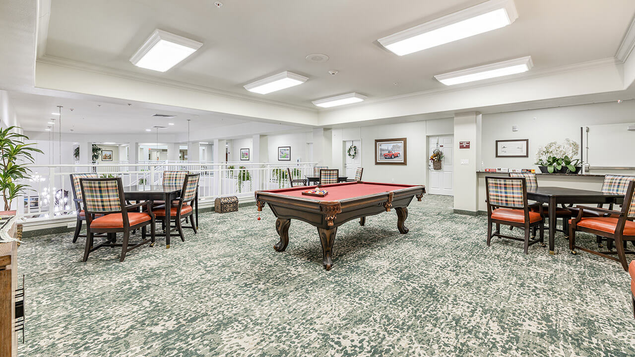 A spacious recreation room with a red-felt pool table at the center, surrounded by tables and plaid-upholstered chairs, bright overhead lights, and framed art on the walls.