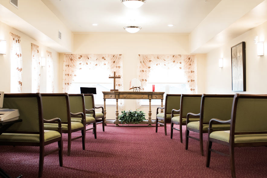 A small chapel with neatly arranged chairs facing a wooden altar. The altar holds a cross, a candle, and a book. The room is well-lit with overhead lights and soft natural light streaming through sheer curtains on the windows behind the altar.