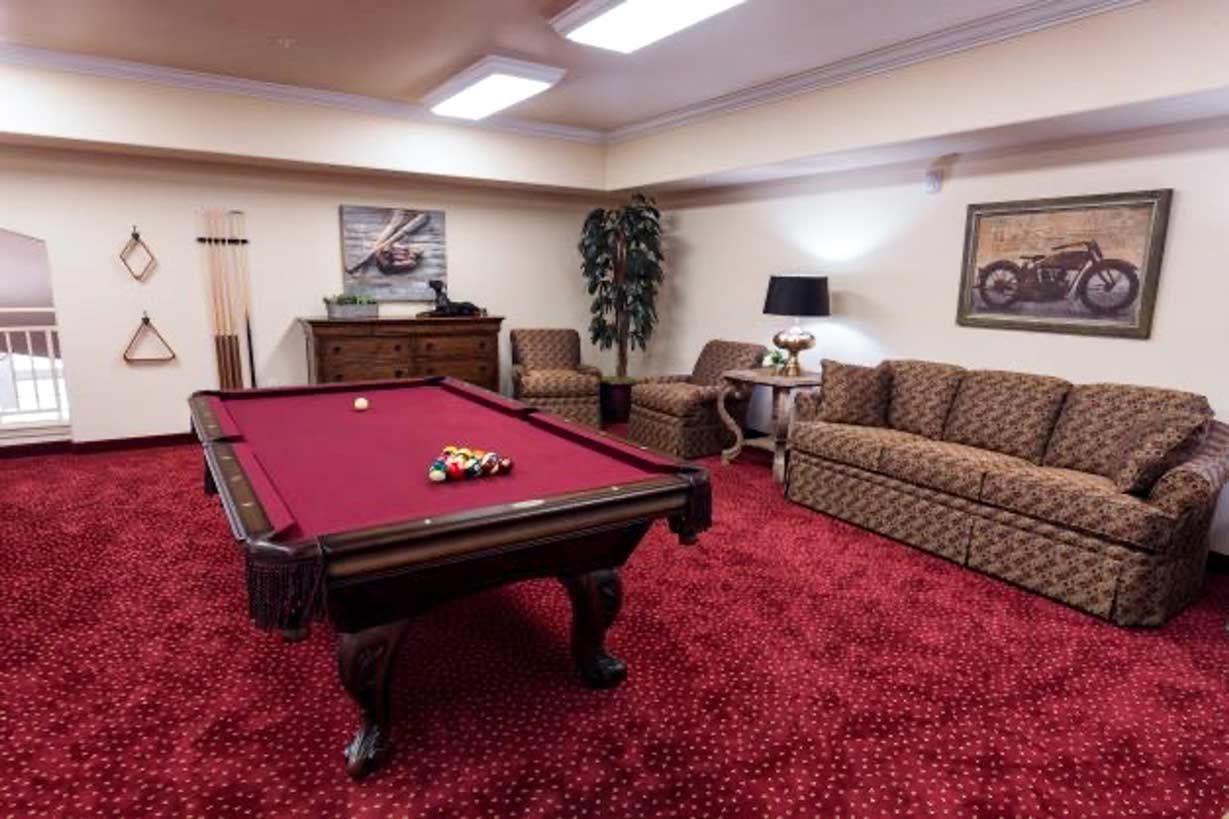 A cozy game room with red carpeted floor featuring a brown pool table with a set of balls arranged at one end. Surrounding furniture includes a floral-patterned sofa, two armchairs, a chest of drawers, and wall decor with car and motorcycle art.